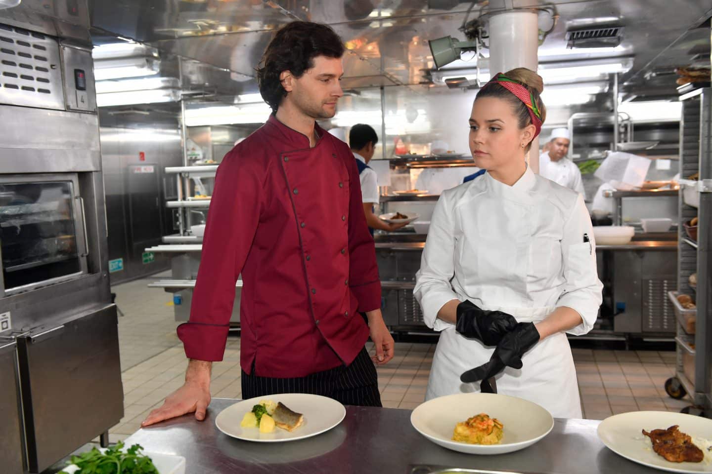 "Das Traumschiff: Argentinien": In der Schiffsküche: Isabell Wagner (Nele Schepe) und Leon Müller (Aaron Karl) stehen nebeneinander vor einer Anrichte, auf der Teller mit diversen Speisen stehen. Während Isabell nachdenklich Handschuhe überstreift, lächelt Leon sie an.
