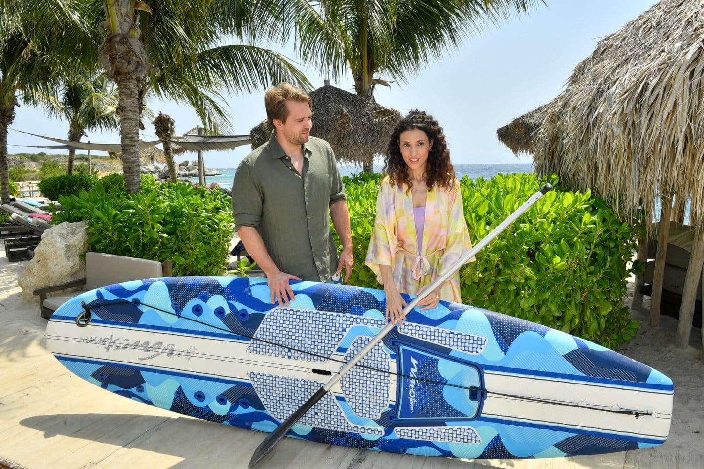 "Das Traumschiff: Curaçao": Am Stand: Valerie Prinz (Nadine Menz) und Philipp Korte (David Korbmann) halten gemeinsam ein Paddleboard. Während Philipp sie anlächelt, blickt Valerie betroffen zu Boden.