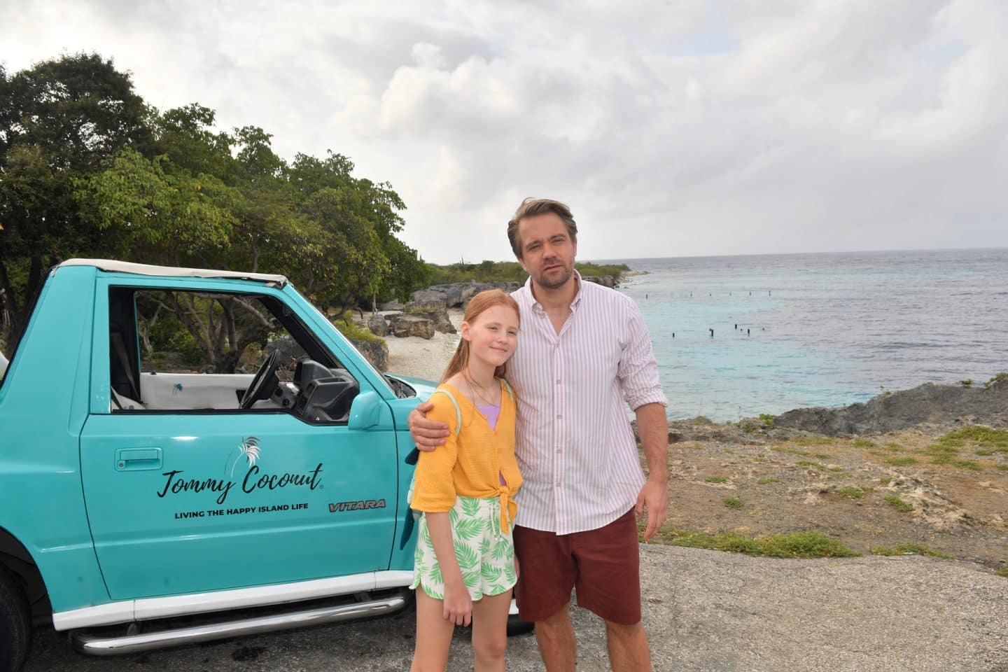 "Das Traumschiff: Curaçao": Auf einem Felsplateau mit Blick auf das Meer: Vor einem Pickup steht Philipp Korte (David Korbmann) und hält seine Tochter Mia (Greta Caspary) im Arm. Beide lächeln.