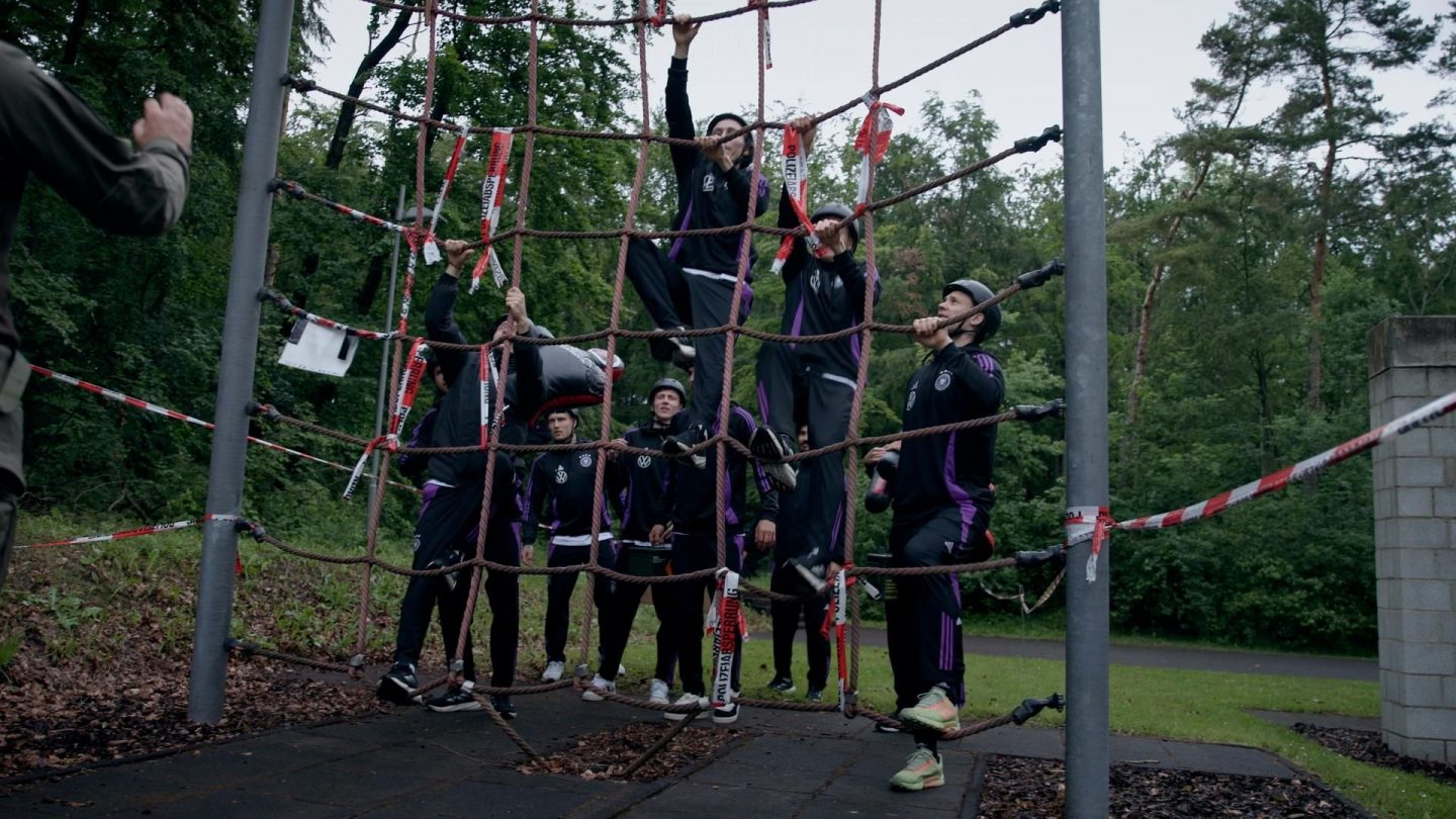 Spieler der deutschen Nationalmannschaft beim Team-Building: Klettern an einem Seilnetz im Freien, begleitet von Anweisungen eines Trainers – eine Übung, die Zusammenhalt und Stärke fördert.