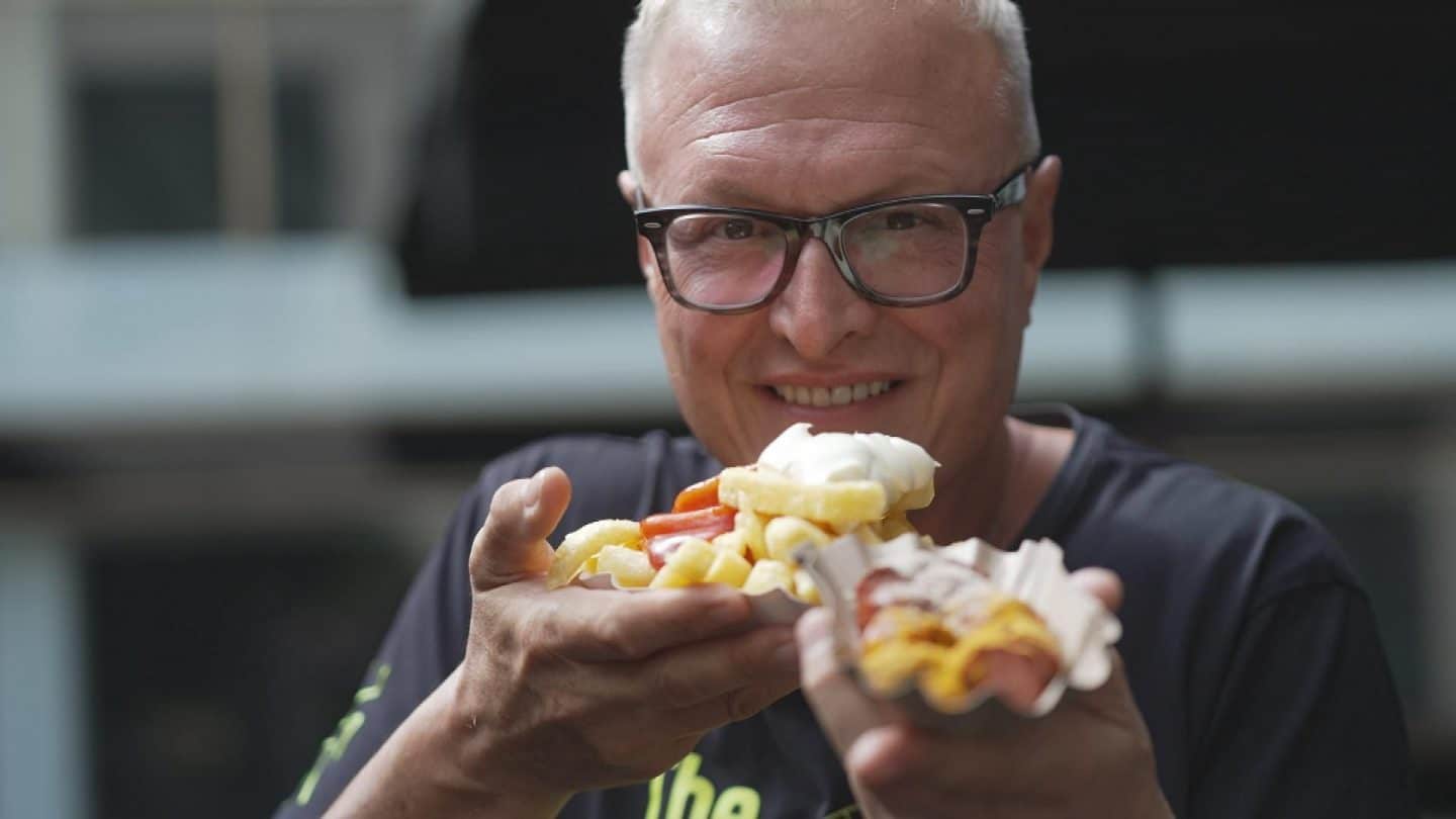 Lars Obendorfer, Geschäftsführer von Best Worscht in Town, lächelt und präsentiert eine Portion Pommes mit Toppings sowie eine Currywurst in einer urbanen Kulisse.