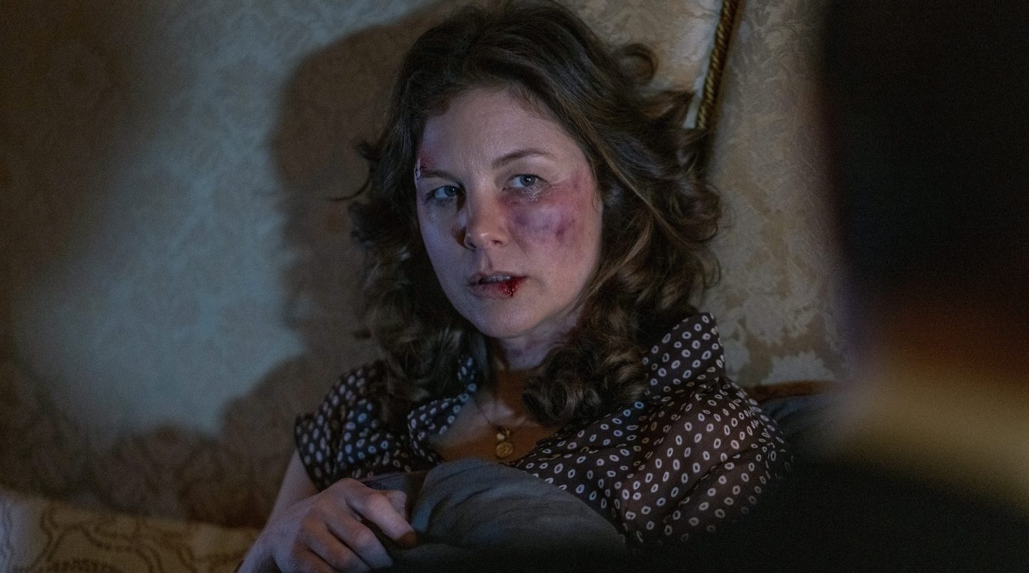 Szene aus „Tatort: Man stirbt nur zweimal“: Eine Frau (Doreen Prätorius, gespielt von Cordelia Wege) mit blauen Flecken und Verletzungen im Gesicht sitzt erschöpft und traumatisiert auf einem Bett. Ihre Haare sind zerzaust, und sie trägt eine gepunktete Bluse. Die düstere Beleuchtung betont die bedrückende und mysteriöse Atmosphäre des Moments.