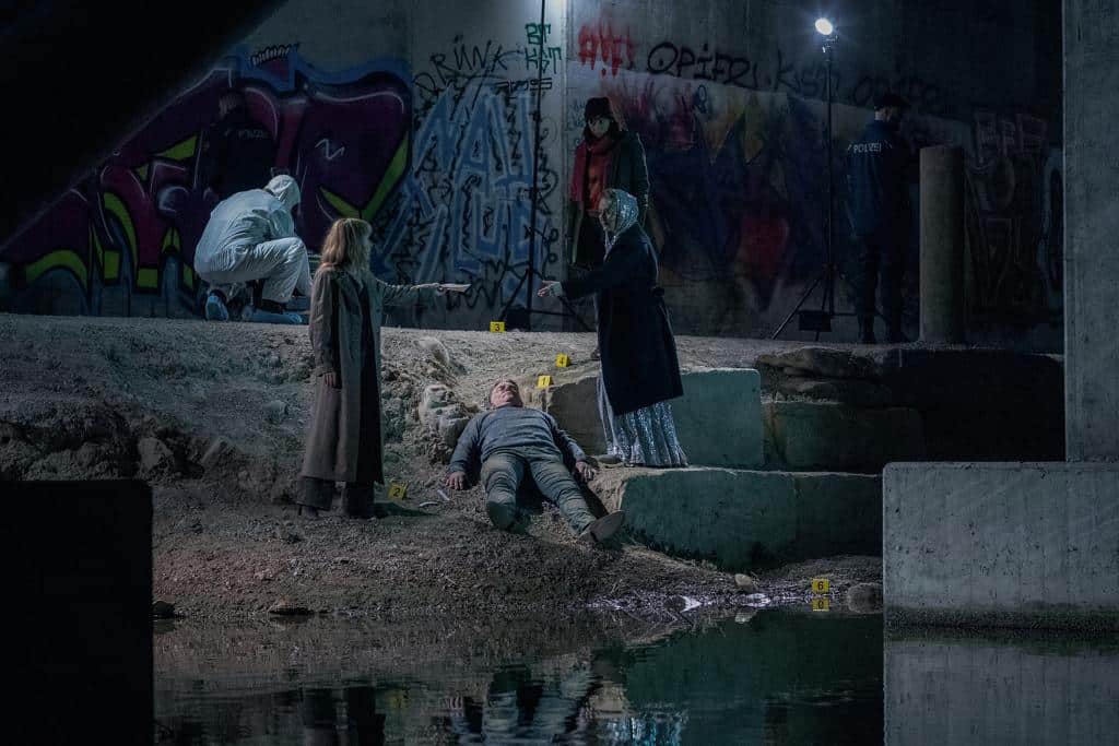 Ein Tatort unter einer Brücke in einer Kanalisation, umgeben von Graffiti. Eine Leiche liegt auf dem Boden, während Kommissarin Isabelle Grandjean (Anna Pieri Zuercher) mit einer Kollegin auf den Tatort deutet. Im Hintergrund sind Kommissarin Tessa Ott (Carol Schuler), Staatsanwältin Anita Wegenast (Rachel Braunschweig) und ein Spurensicherungsteam bei der Arbeit zu sehen. Die Szene ist düster und mit Lichtspots ausgeleuchtet.