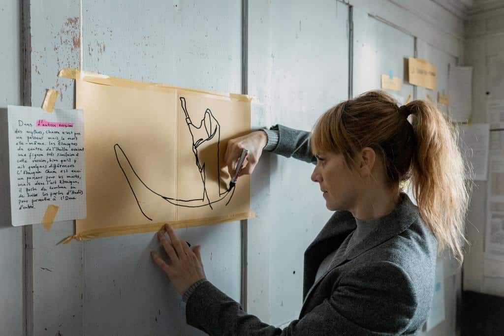 Kommissarin Isabelle Grandjean (Anna Pieri Zuercher) analysiert an einer Wand eine Zeichnung, die einen Fährmann in einem Boot darstellt. Neben der Skizze hängen handschriftliche Notizen, die an die Wand geklebt wurden. Grandjean wirkt konzentriert, während sie Details untersucht.