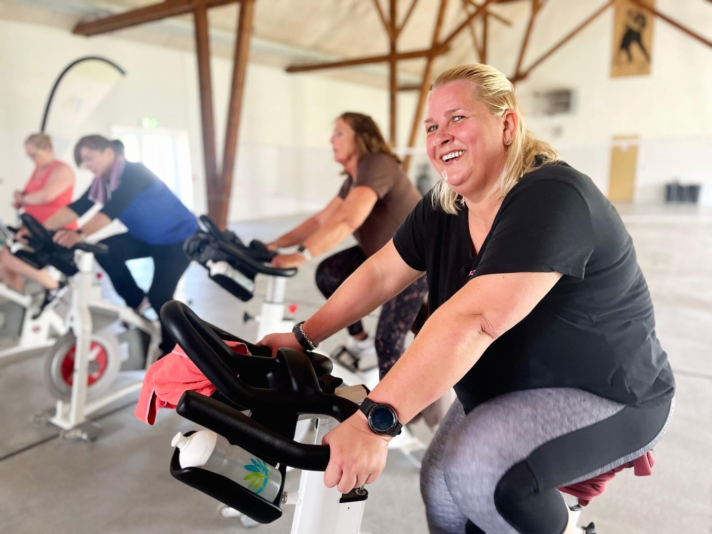 Bianca in Sportkleidung lächelt, während sie auf einem Indoor-Fahrrad trainiert. Im Hintergrund trainieren weitere Personen in einer hellen Sporthalle mit Holzbalken