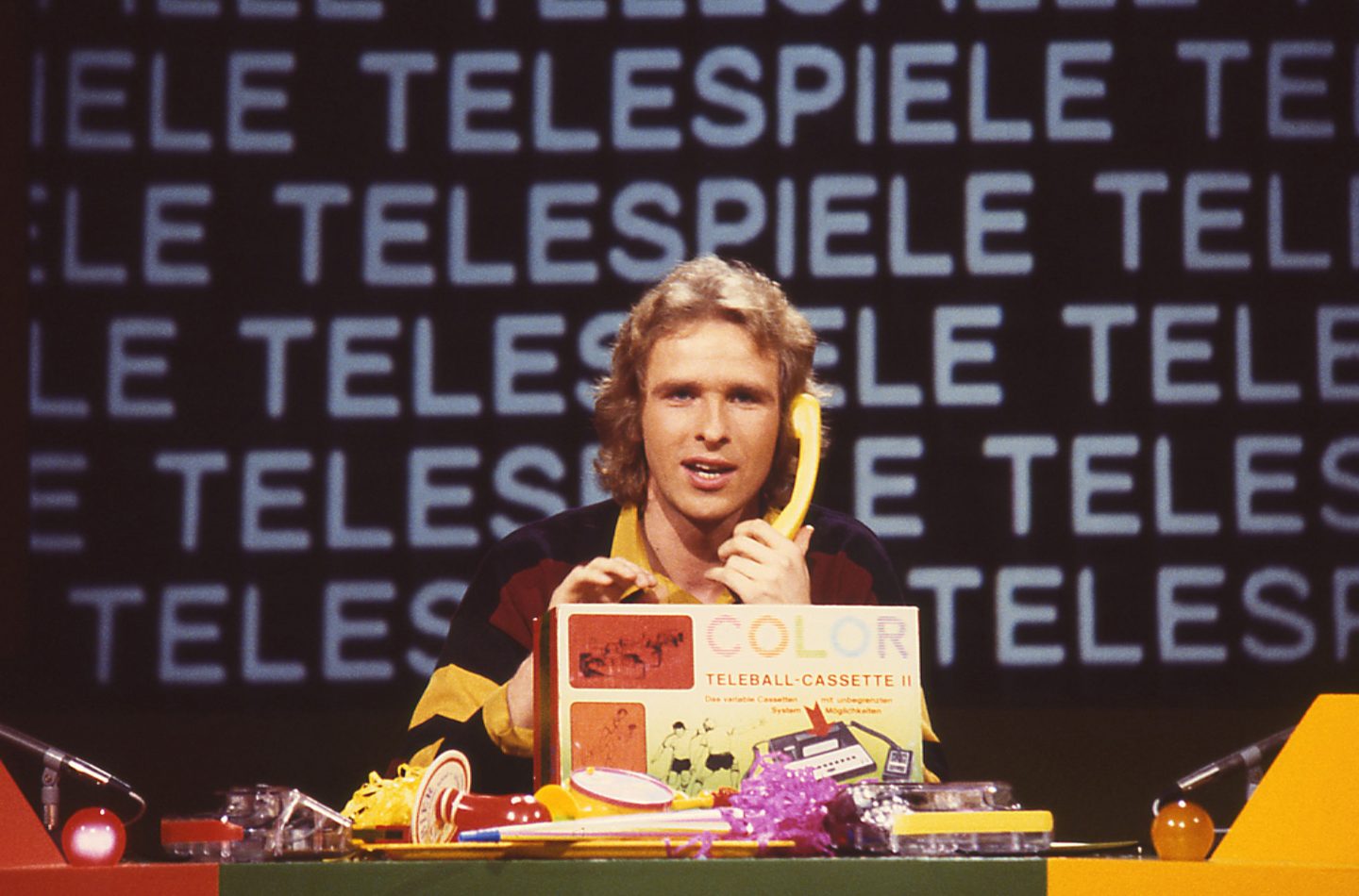Thomas Gottschalk moderiert die Kult-Spielshow "Telespiele", die zwischen 1977 und 1981 in 29 Folgen ausgestrahlt wurde. Im Bild sitzt er mit einem gelben Telefon vor Requisiten, darunter eine "Teleball-Cassette II". Die Show war Gottschalks erste Live-TV-Sendung und markierte den Beginn seiner Fernsehkarriere. Im Hintergrund ist das Wort "Telespiele" mehrfach zu sehen.