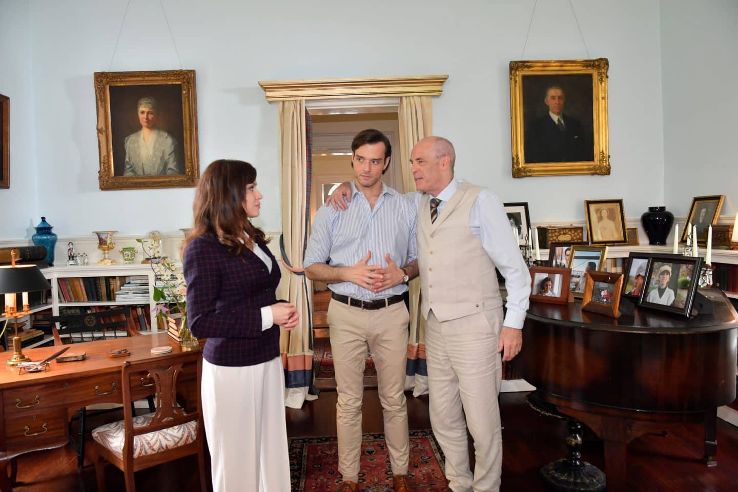 Das Traumschiff: Hudson Valley": Im Wohnraum der Villa: Stolz hat Hendrik Hansen (Peter Kremer) einen Arm um seinen Sohn Jasper Hansen (Marc Schöttner) gelegt. Dieser blickt fragend seine Schwester Frederike (Natalie Avelon) an, die ihnen gegenüber steht. Der Raum ist vollgestellt mit Familienfotos.