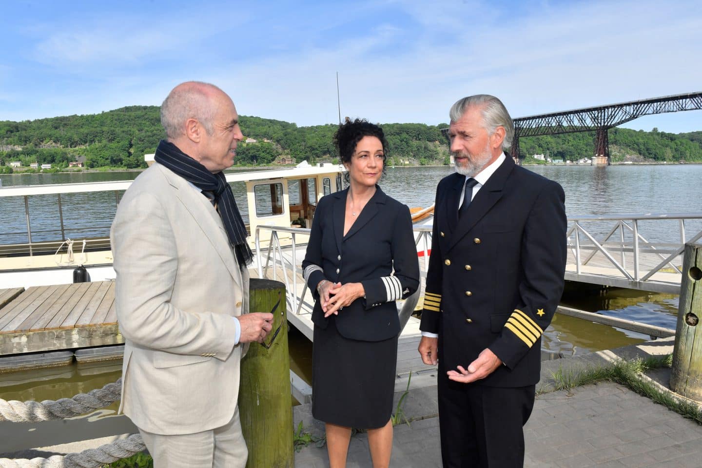 "Das Traumschiff: Hudson Valley": Vor einem Bootsanleger: Mit erklärender Gestik steht Staff-Kapitän Martin Grimm (Daniel Morgenroth) vor Hendrik Hansen (Peter Kremer). Hanna Liebhold (Barbara Wussow) steht neben ihm und blickt Martin unterstützend an.
