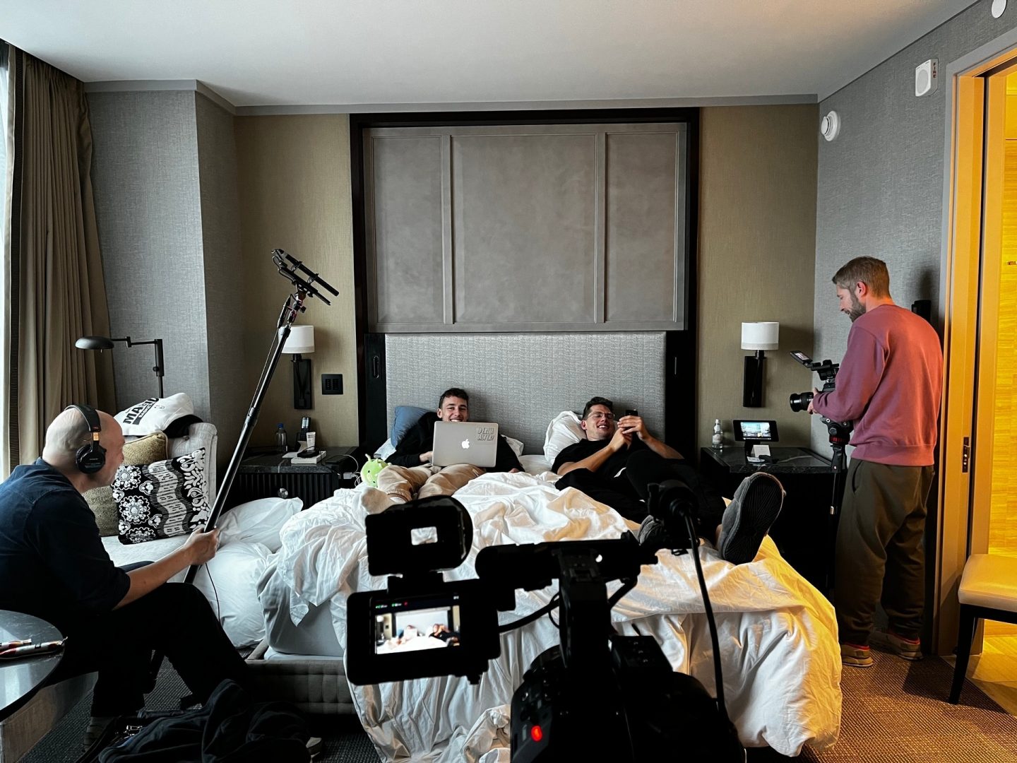 Die Brüder Wagner liegen mit Laptop und Smarphone in der Hand liegen mit Straßenkleidung in einem Hotelbett, während ein Mann mit Fotoapparat neben dem Bett steht und ein Mann mit Kopfhörer und Tonangel mit Mikrofon vor dem Fenster sitzt.
