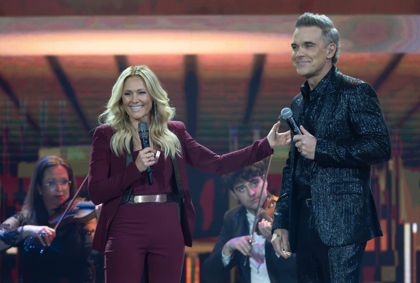 Die Helene Fischer-Show": Helene Fischer (l.) und Robbie Williams (r.) stehen nebeneinander auf der Bühne - im Hintergrund zwei ViolinistInnen. Helene Fischer trägt einen weinroten Hosenanzug mit einem silbernen Gürtel, sie berührt Robbie Williams mit ihrer linken Hand an seiner rechten Schulter und blickt lächelnd nach links aus dem Bild. Robbie Williams trägt einen glitzernden, schwarzen Anzug. Auch er lächelt und schaut nach links. Beide halten je ein Mikrofon in der Hand. Die Kameraeinstellungsgröße ist amerikanisch.
