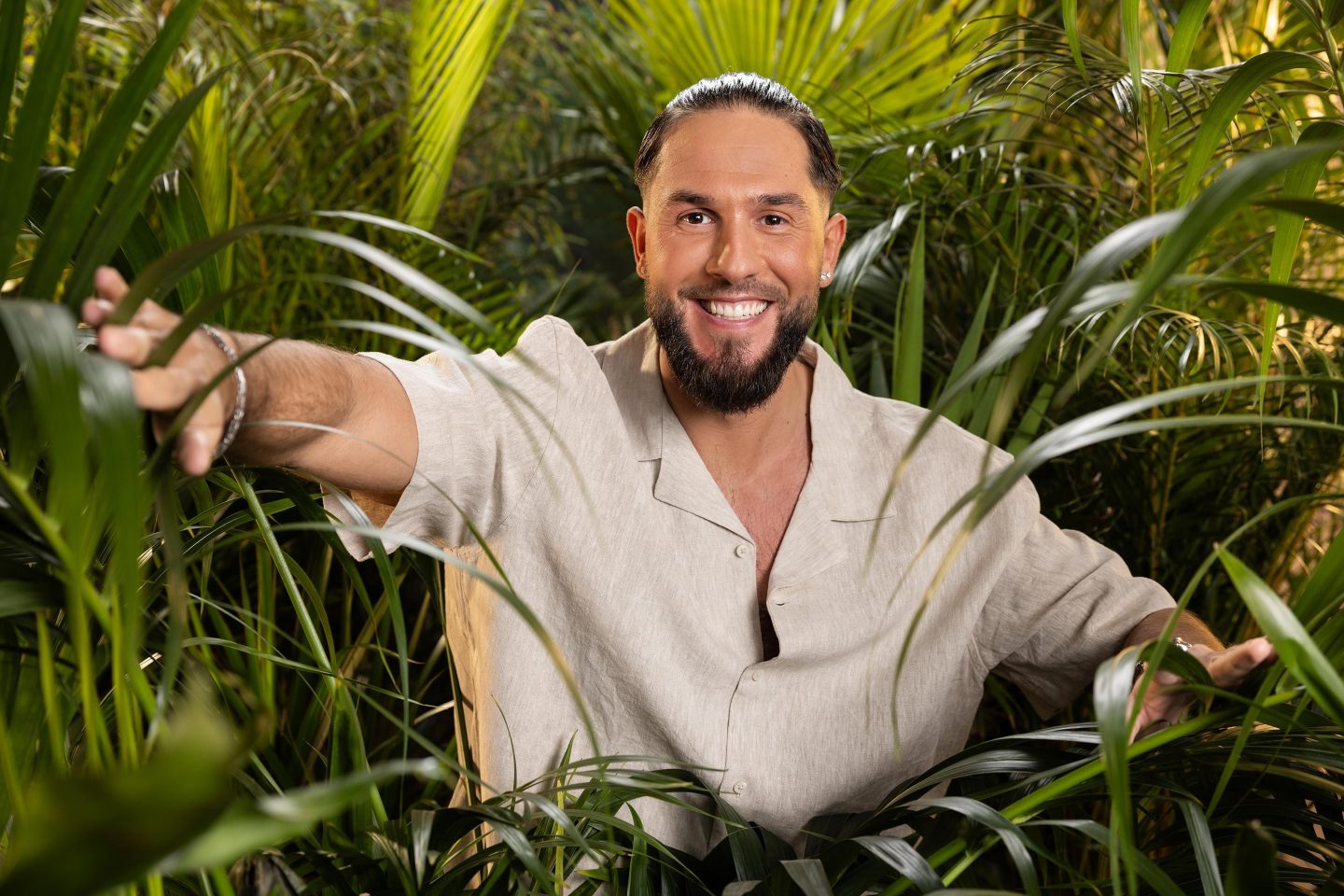 Maurice Dziwak steht lächelnd inmitten von tropischen Pflanzen. Er trägt ein helles, lässiges Hemd und streckt seine Arme aus, während er fröhlich in die Kamera schaut. Die grüne Dschungelkulisse unterstreicht das Abenteuer-Thema.