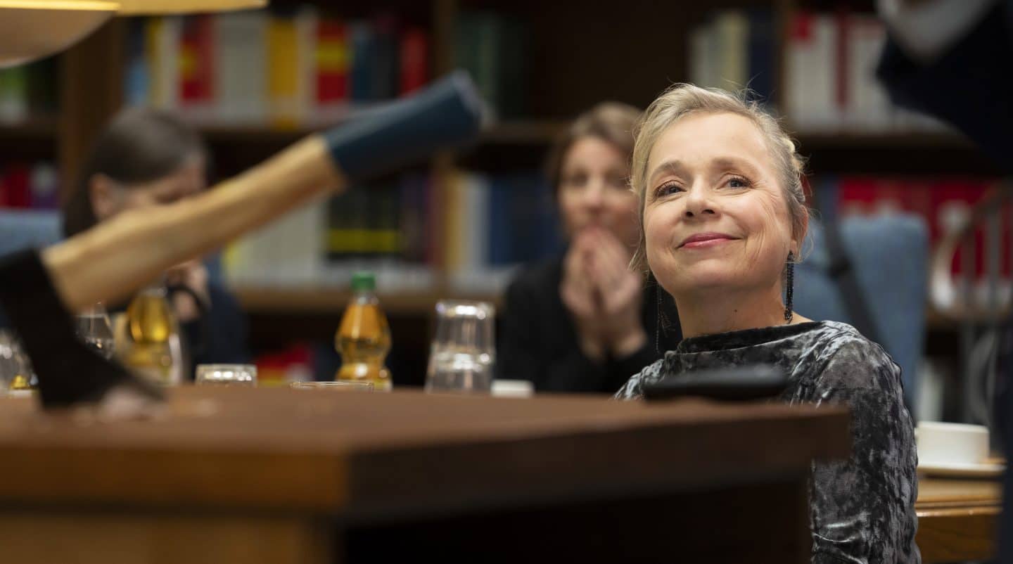 Szene aus „Tatort: Man stirbt nur zweimal“: Eine Frau (Doreen Prätorius, gespielt von Cordelia Wege) sitzt lächelnd in einem Raum voller Bücherregale. Im Vordergrund liegt eine Axt auf einem Tisch, die verschwommen zu sehen ist. Die Frau trägt ein graues, gemustertes Oberteil, während sie von anderen Personen im Hintergrund beobachtet wird. Die Atmosphäre wirkt angespannt und rätselhaft.