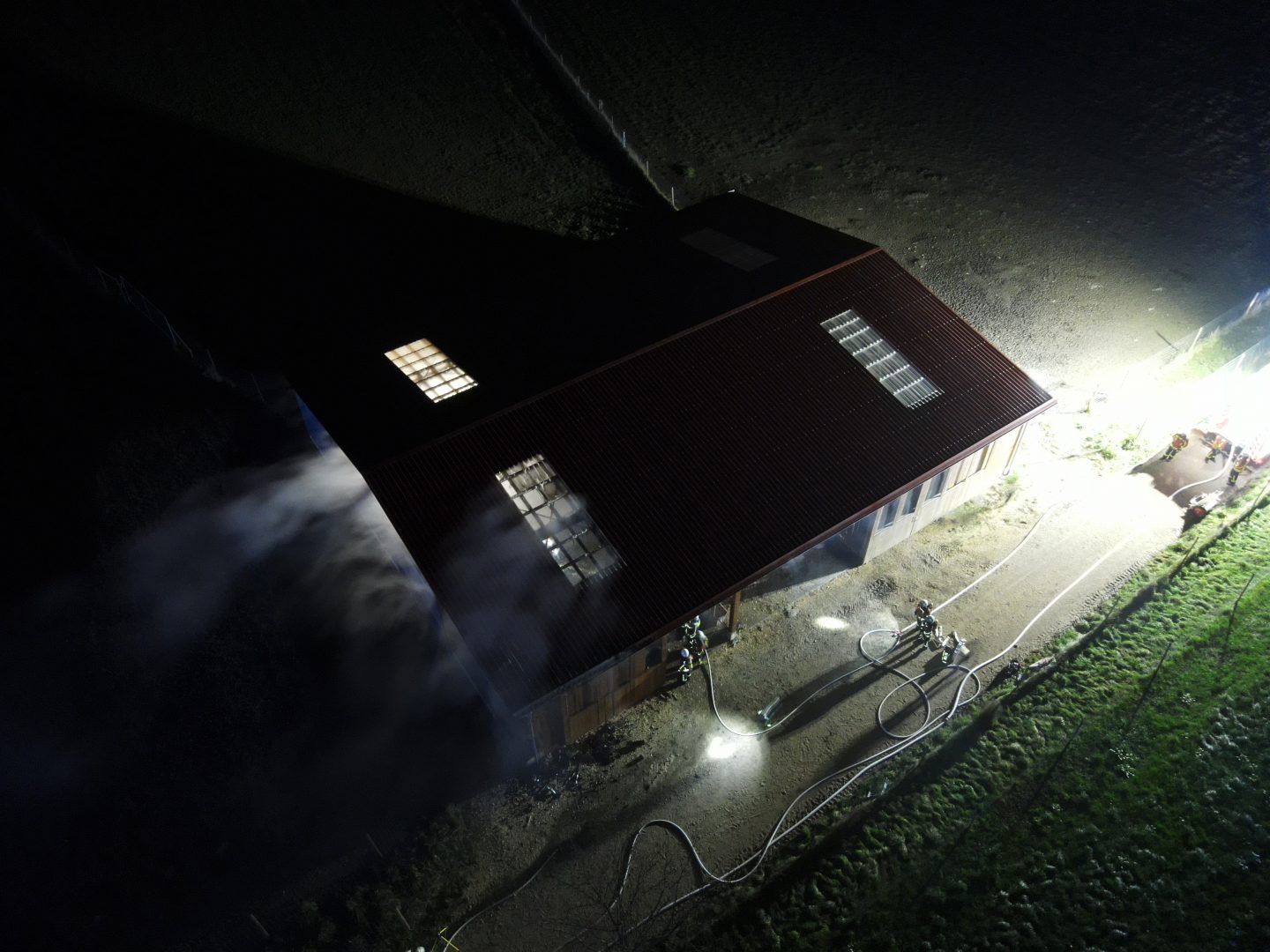 Luftaufnahme des Stallgebäudes in Stockach-Airach während der Löscharbeiten bei Nacht. Rauch steigt aus dem Dach, während Feuerwehrkräfte die Umgebung absichern.