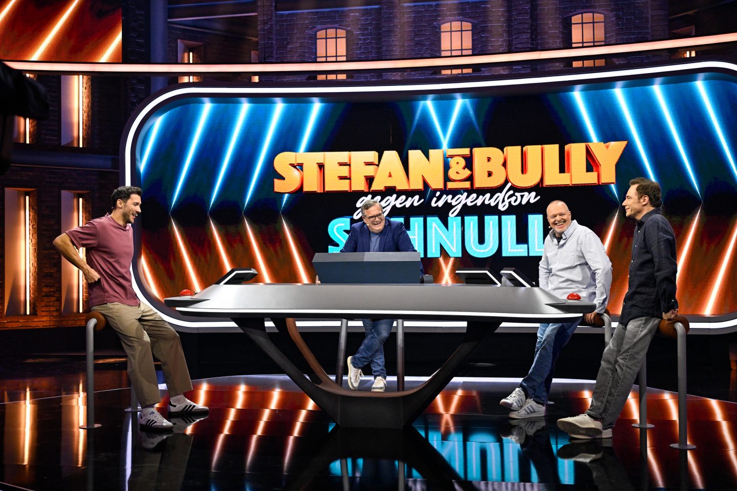 V.l.: Kandidat Marc, Moderator Elton, Stefan Raab und Michael 'Bully' Herbig stehen gemeinsam an einem modernen Studiopult in der Show 'Stefan & Bully gegen irgendson Schnulli', im Hintergrund das beleuchtete Show-Logo.