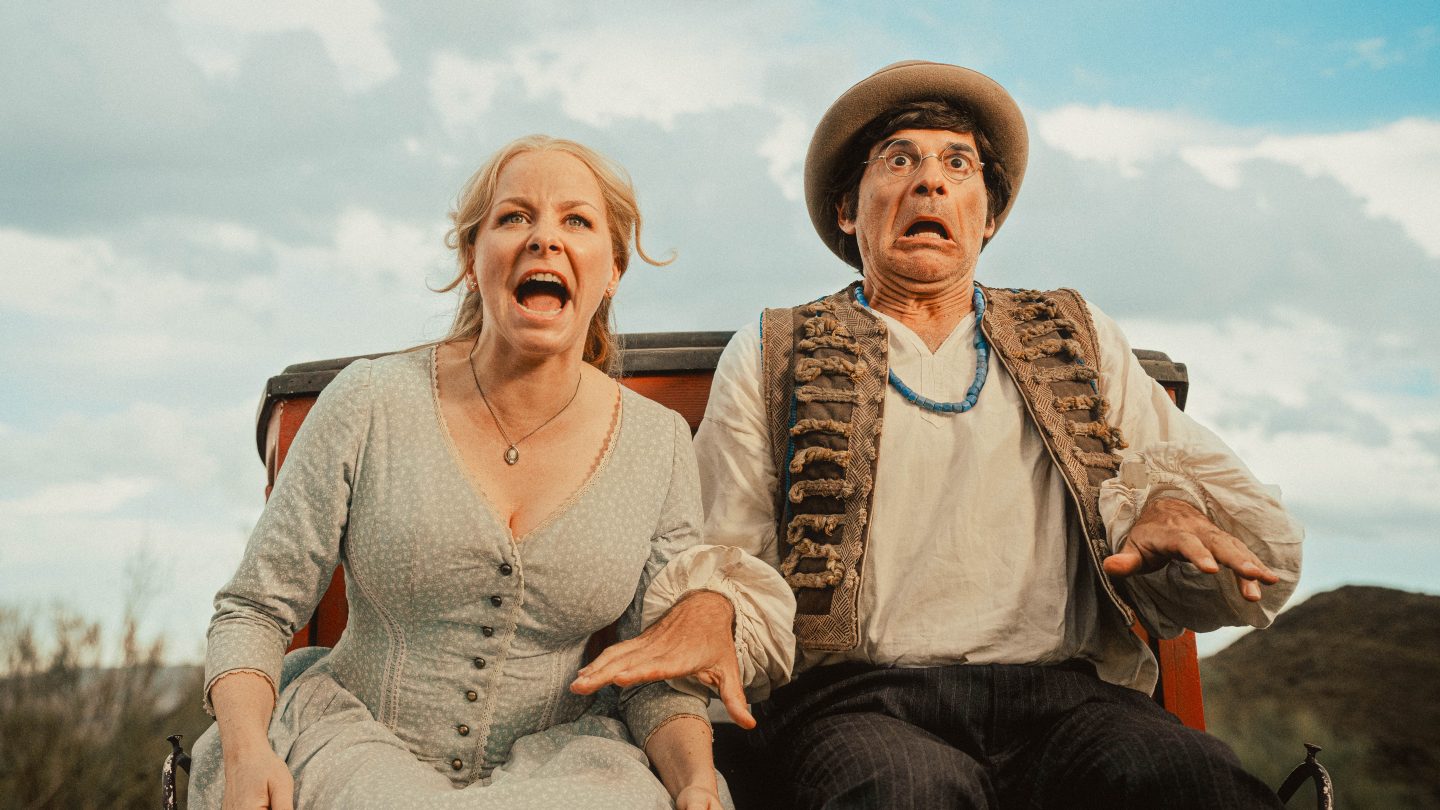 Jasmin Schwiers als Mary und Rick Kavanian als Dimitri sitzen in einer Pferdekutsche und zeigen erschrockene Gesichter. Mary trägt ein helles Kleid, während Dimitri mit Weste, Perlenkette und Hut bekleidet ist. Der Hintergrund zeigt eine Wildwestlandschaft mit blauem Himmel.