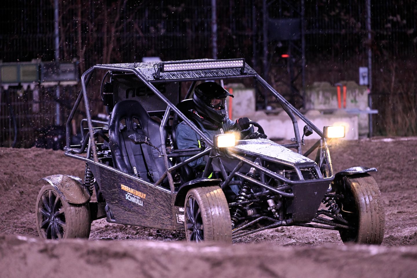 Ein Fahrer in Schutzkleidung und Helm steuert einen Offroad-Buggy mit dem Schriftzug 'Stefan & Bully Schnulli' durch eine schlammige Rennstrecke bei Nacht, während helle Scheinwerfer den Weg erleuchten.