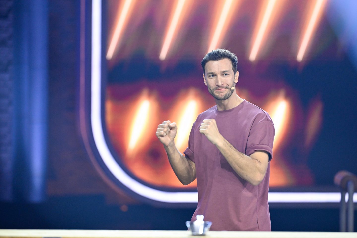 Marc, der Kandidat der neuen RTL-Show 'Raab und Bully gegen irgendson Schnulli', steht siegessicher mit erhobenen Fäusten auf der Bühne, im Hintergrund ein leuchtendes Bühnendesign in Orange- und Rottönen.