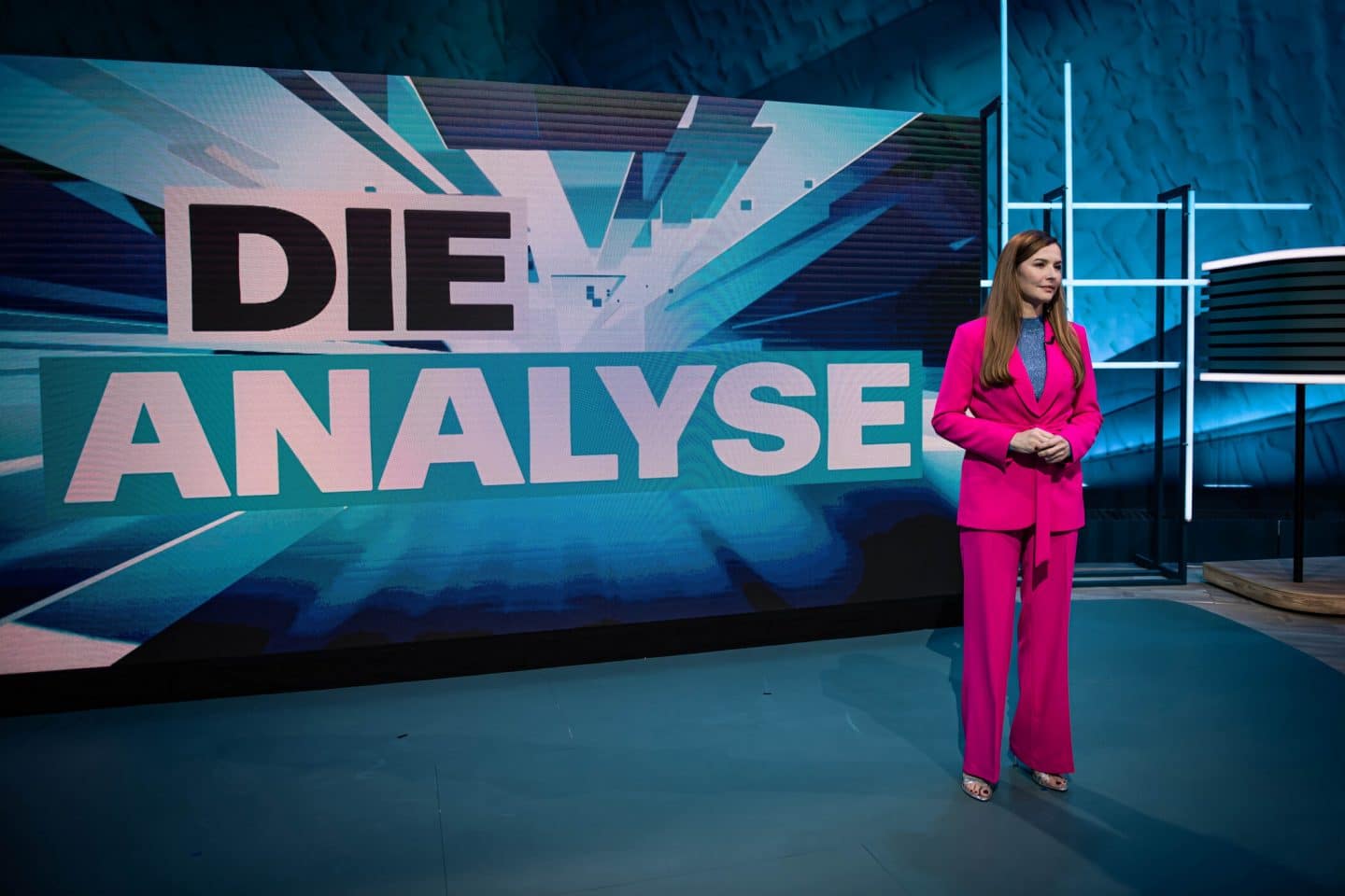 Linda Zervakis im auffälligen pinken Hosenanzug steht vor einem großen Bildschirm im Studio. Auf dem Bildschirm prangt der Schriftzug „DIE ANALYSE“ in großen Buchstaben. Das Setting ist modern und in blauen Farbtönen gehalten.