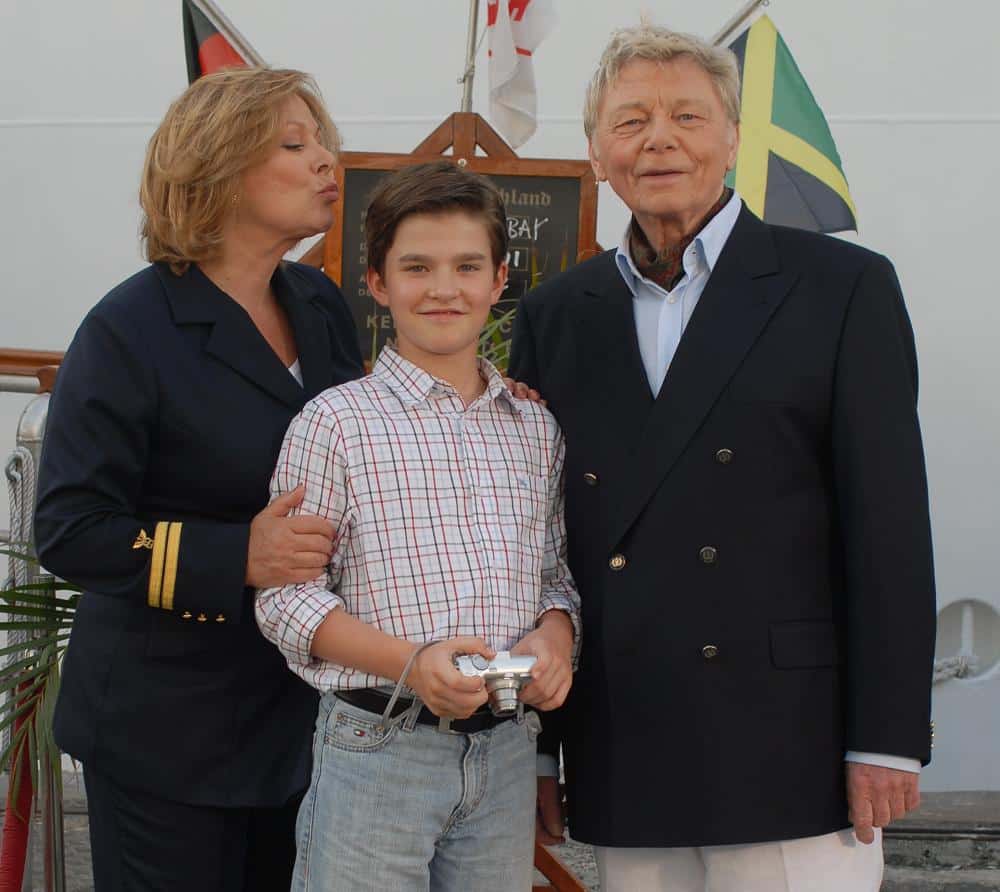 Eine Szene hinter den Kulissen der Dreharbeiten zur Traumschiff-Folge 'San Francisco' (2007): Heide Keller in Uniform lächelt neben Patrick Mölleken (Mitte), der eine Kamera in der Hand hält, und Uwe Friedrichsen (rechts) in einem eleganten Doppelreiher. Im Hintergrund sind Flaggen und eine Tafel mit der Aufschrift 'Jamaica' zu sehen.