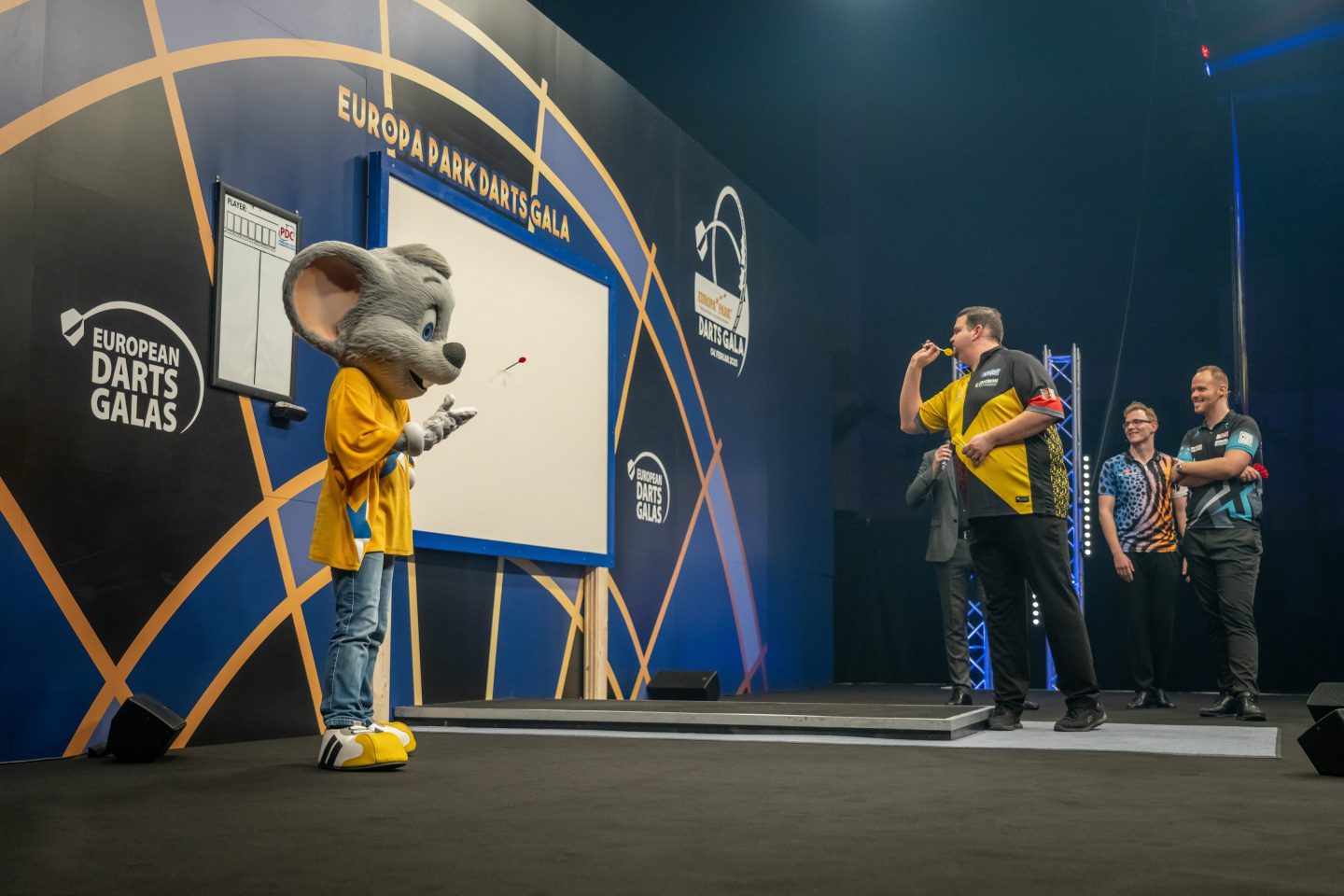 Gabriel Clemens, bekannt als 'German Giant', tritt auf der Bühne der Europa-Park Arena in Rust bei der PDC Darts Gala auf. Neben ihm sind weitere Darts-Stars und das Maskottchen des Europa-Parks zu sehen, während die Zuschauer die Action verfolgen.