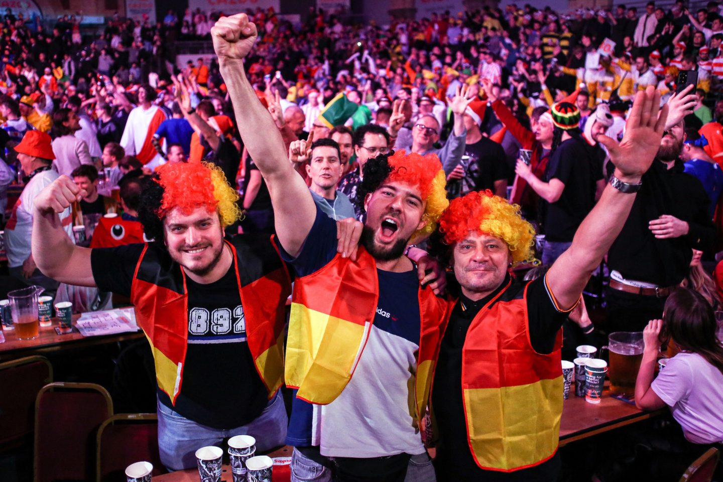 Begeisterte Darts-Fans in der Europa-Park Arena: Drei Männer in Schwarz-Rot-Gold gekleidet, mit Perücken in den Farben der deutschen Flagge, jubeln ausgelassen inmitten einer feiernden Menge.