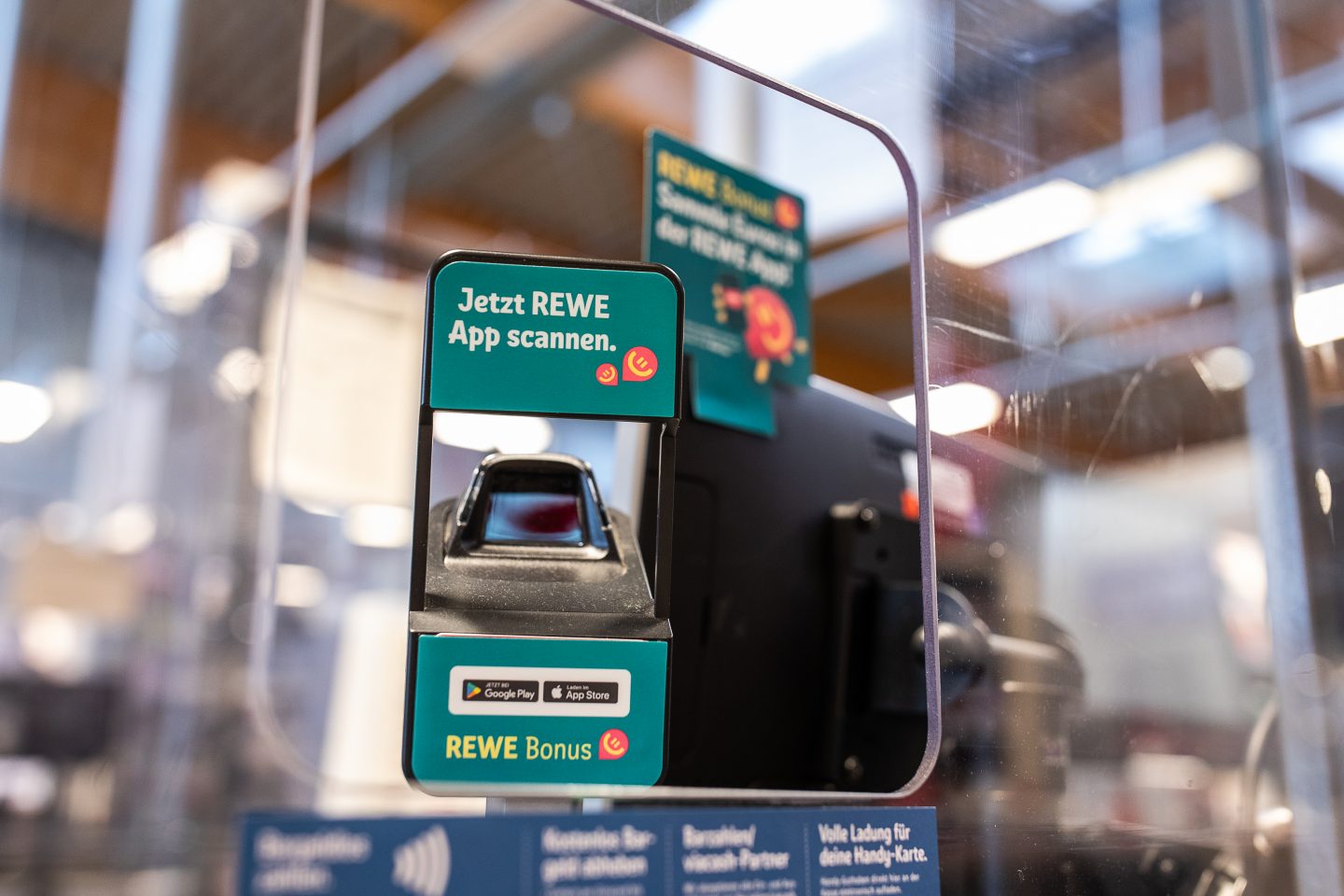 Ein Scanner an der Kasse eines REWE-Marktes mit Hinweisen auf das neue REWE Bonusprogramm. Kunden können die REWE App scannen, um Euro-Guthaben zu sammeln und direkt einzulösen. Im Hintergrund sind weitere Hinweise auf das Bonusprogramm sichtbar.