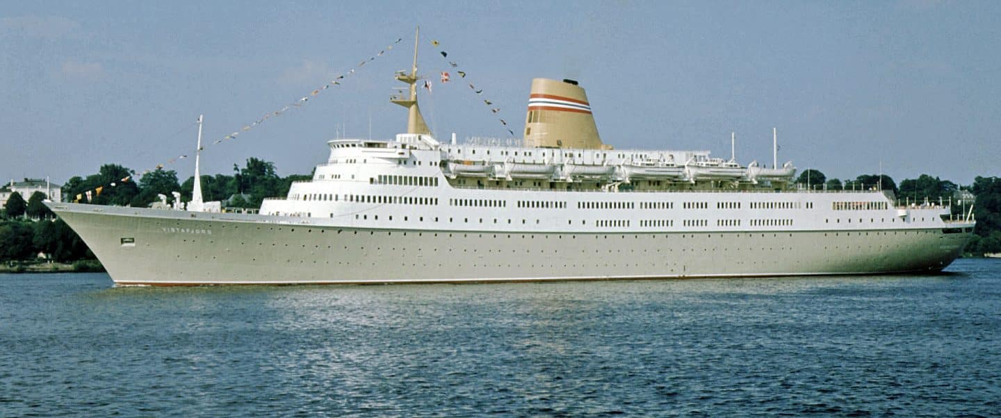 Ein großes, elegantes Kreuzfahrtschiff, die norwegische Vistafjord, auf ruhigem Wasser, geschmückt mit Flaggen auf der Mastspitze und einer weißen, klassischen Lackierung. Das Schiff war eines der ersten Traumschiffe.