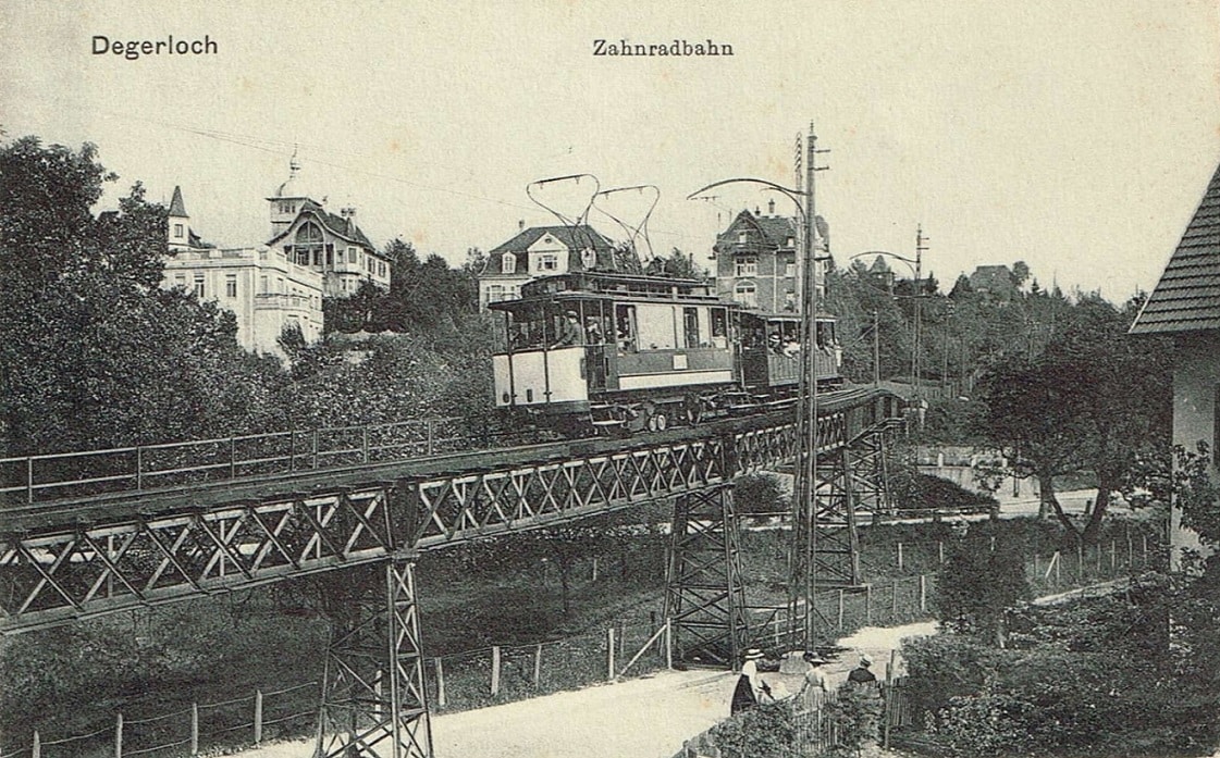 Ein Triebwagen der ersten elektrischen Generation der Stuttgarter Zahnradbahn fährt mit einem Vorstellwagen bergauf über eine Stahlbrücke in der Alten Weinsteige. Im Hintergrund sind die historischen Villen der Degerlocher Villenkolonie zu sehen.






