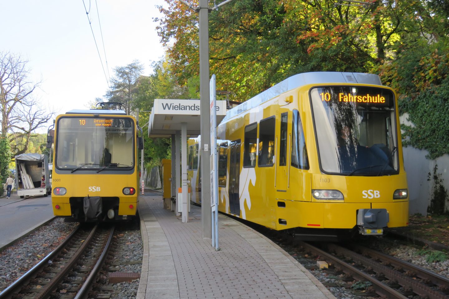 Zwei gelbe Triebwagen der Stuttgarter Zahnradbahn stehen an der Haltestelle Wielandshöhe nebeneinander. Links ist ein Fahrzeug der dritten Generation (ZT 4), rechts ein moderner Triebwagen der vierten Generation (ZT 4.2) mit der Aufschrift „Fahrschule“ zu sehen. Die Szene wird von herbstlichem Laub und der charakteristischen Architektur der Haltestelle umrahmt.






