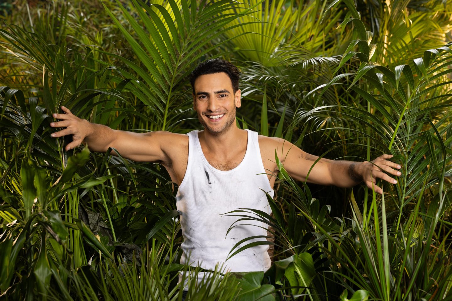 Timur Ülker, lächelnd in einem weißen, leicht verschmutzten Tanktop, mit ausgestreckten Armen zwischen tropischen Pflanzen im Dschungel stehend.







