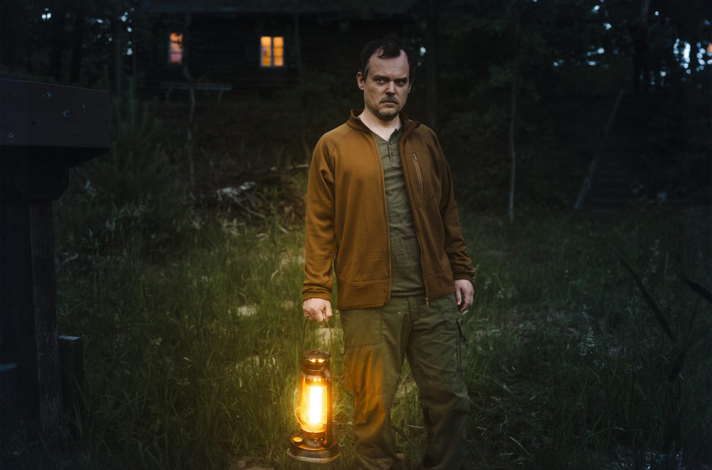 Olaf Brandstädt (Johannes Allmayer) steht nachts vor seiner Anglerhütte und hält eine leuchtende Petroleumlampe in der Hand. Sein Blick ist ernst und misstrauisch, während im Hintergrund die schwach beleuchtete Hütte in der Dunkelheit verschwindet. Die Szene wirkt geheimnisvoll und bedrohlich, mit dunklem Wald und hohem Gras, das die Atmosphäre verstärkt.