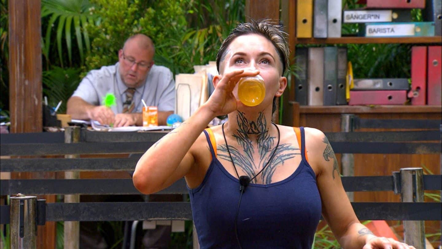 Edith Stehfest während der Dschungelprüfung „Bährufs-Bähratung“, trinkt ein Glas mit gelber Flüssigkeit, während im Hintergrund ein Mitarbeiter die Situation beobachtet.