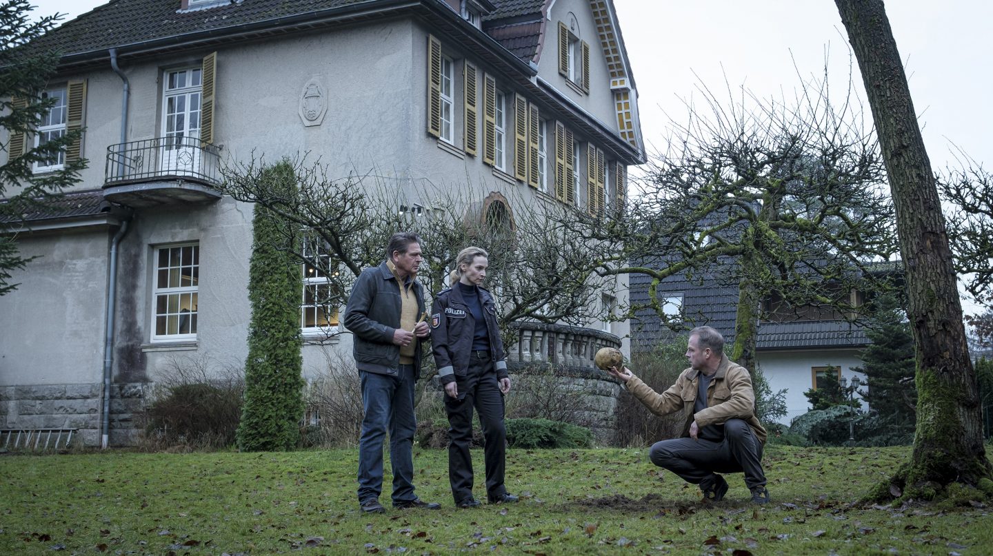 Vor dem Nolden-Haus stehen Puttkammer (gespielt von Joshy Peters), Hannah Wagner (gespielt von Jana Klinge) und Hauke Jacobs (gespielt von Hinnerk Schönemann). Hauke kniet im Gras und hält einen Schädel in der Hand, während die anderen beiden besorgt zusehen. Die Szene deutet auf die Entdeckung eines lange vergrabenen Geheimnisses hin und vermittelt eine beklemmende, geheimnisvolle Atmosphäre.