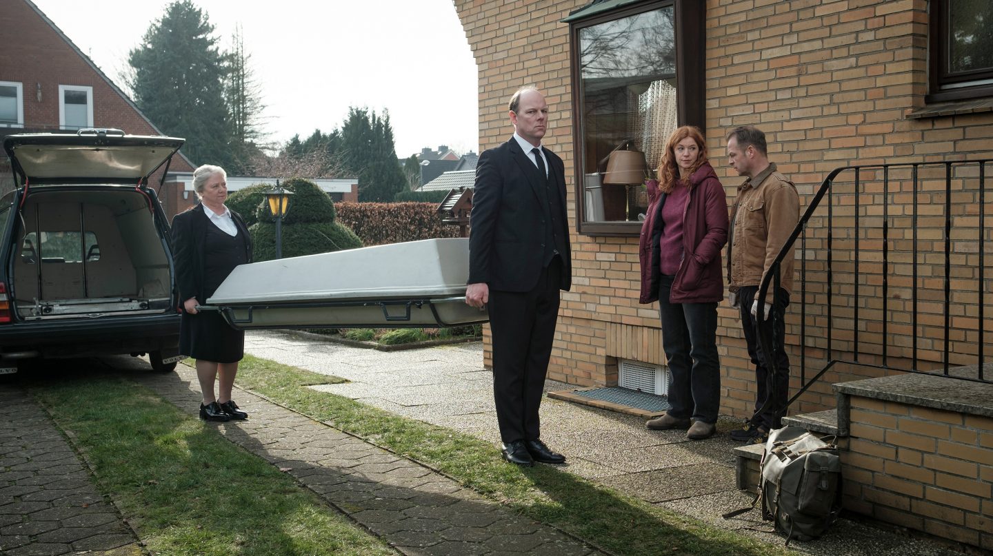 Frau Bleckmann (Regine Hentschel) und Herr Töteberg (Stephan A. Tölle) transportieren eine Trage vor einem Wohnhaus, während zwei Personen, darunter Hauke Jacobs (Hinnerk Schönemann), beobachtend daneben stehen.