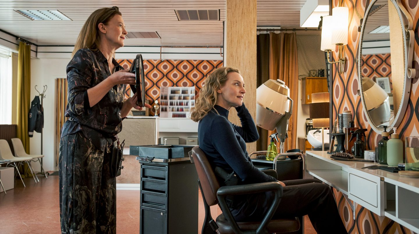 Hannah Wagner (Jana Klinge) sitzt lächelnd auf einem Friseurstuhl, während Grit Hartmann (Julika Jenkins) mit einem Spiegel in ihrem Retro-Salon aus den 70er-Jahren steht.