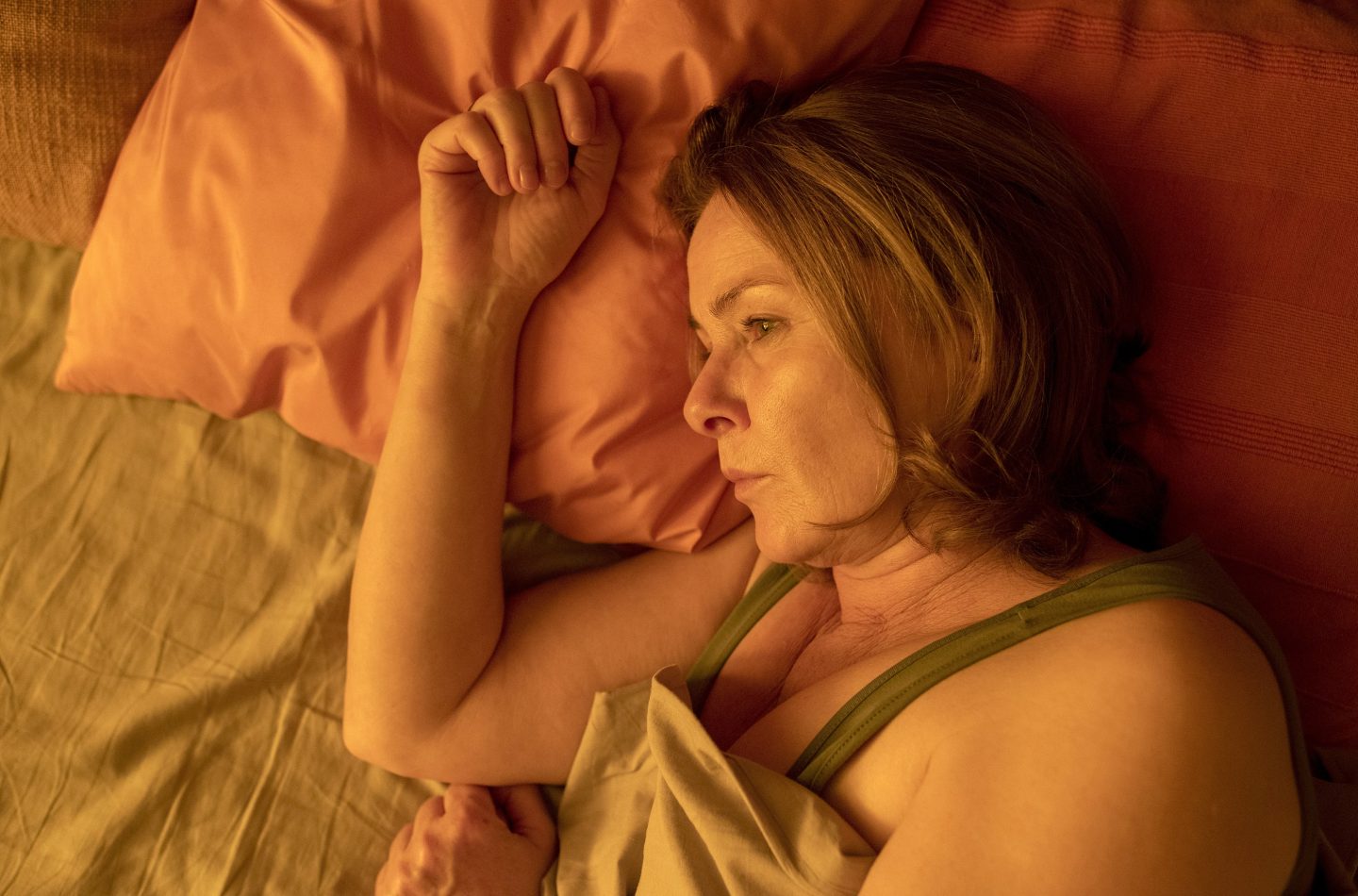 Die Schauspielerin Lina Wendel als Barbara Döring liegt nachdenklich in einem Bett, umgeben von warmem, gedämpftem Licht. Ihre Kopfhaltung und der Ausdruck wirken melancholisch und introspektiv, während sie auf einem orangen Kissen ruht