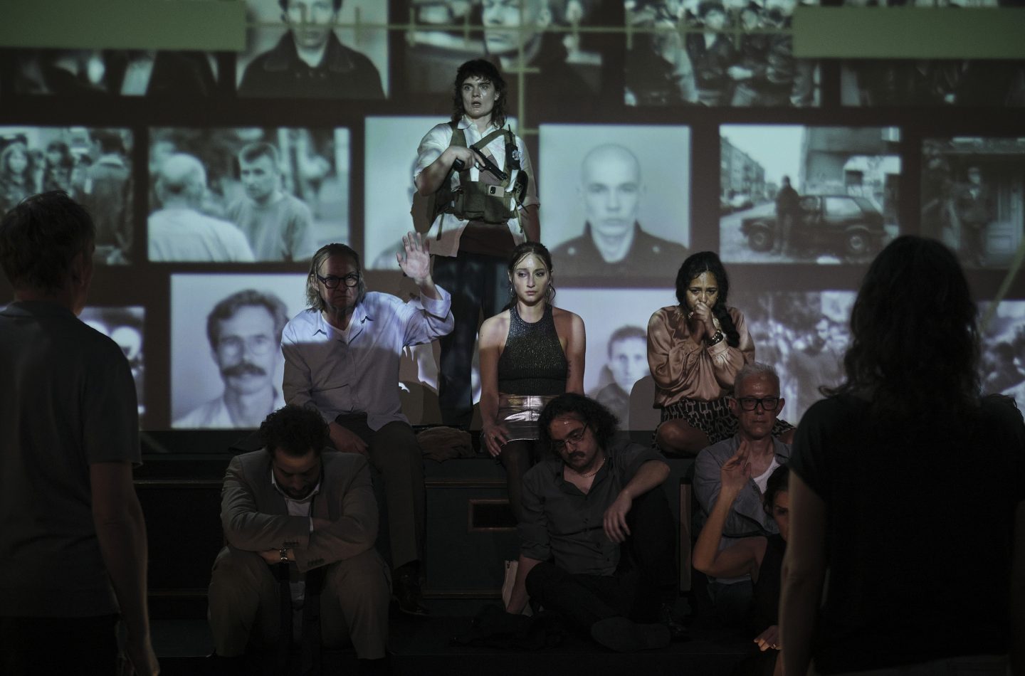 Eine bewaffnete Geiselnehmerin steht vor einer Wand mit projizierten Bildern von Personen. Vor ihr sitzen mehrere Geiseln auf einer Stufe, sichtlich verängstigt. Ein Mann in einem weißen Hemd hebt zögerlich die Hand, während andere Geiseln mit besorgten und verzweifelten Gesichtsausdrücken auf den Boden oder in die Ferne blicken. Die düstere Beleuchtung und die angespannte Körpersprache der Personen unterstreichen die bedrohliche Atmosphäre der Situation.