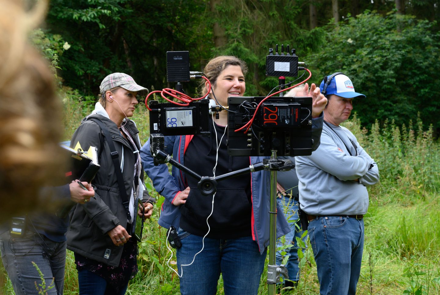 Regisseurin Tini Tüllmann steht lächelnd hinter einem professionellen Monitor-Setup während der Dreharbeiten im Freien. Sie trägt eine dunkelblaue Jacke mit roten Akzenten und hat Kopfhörer im Ohr. Neben ihr steht eine Frau mit einer Kappe und einer grauen Jacke, die konzentriert auf ein Klemmbrett schaut. Rechts im Bild steht ein Mann mit blauer Kappe und Kopfhörern, der mit verschränkten Armen das Geschehen beobachtet. Im Hintergrund ist eine grüne Waldlandschaft zu sehen, die den Drehort umgibt.