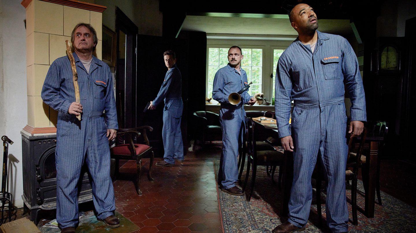 In einem Raum mit rustikaler Einrichtung stehen vier Männer in identischen blauen Overalls. Kaminski (gespielt von V. Leittersdodrf) hält einen Stock in der Hand, während Meyer (gespielt von K. Lindhorst) mit einer Waffe in der Tür steht. Gericke (gespielt von R. Knižka) trägt ein Horn, und Dudek (gespielt von T. Führer) schaut nach oben, als ob er etwas im Raum wahrnimmt. Die angespannte Haltung der Männer deutet auf eine Flucht oder eine brenzlige Situation hin.