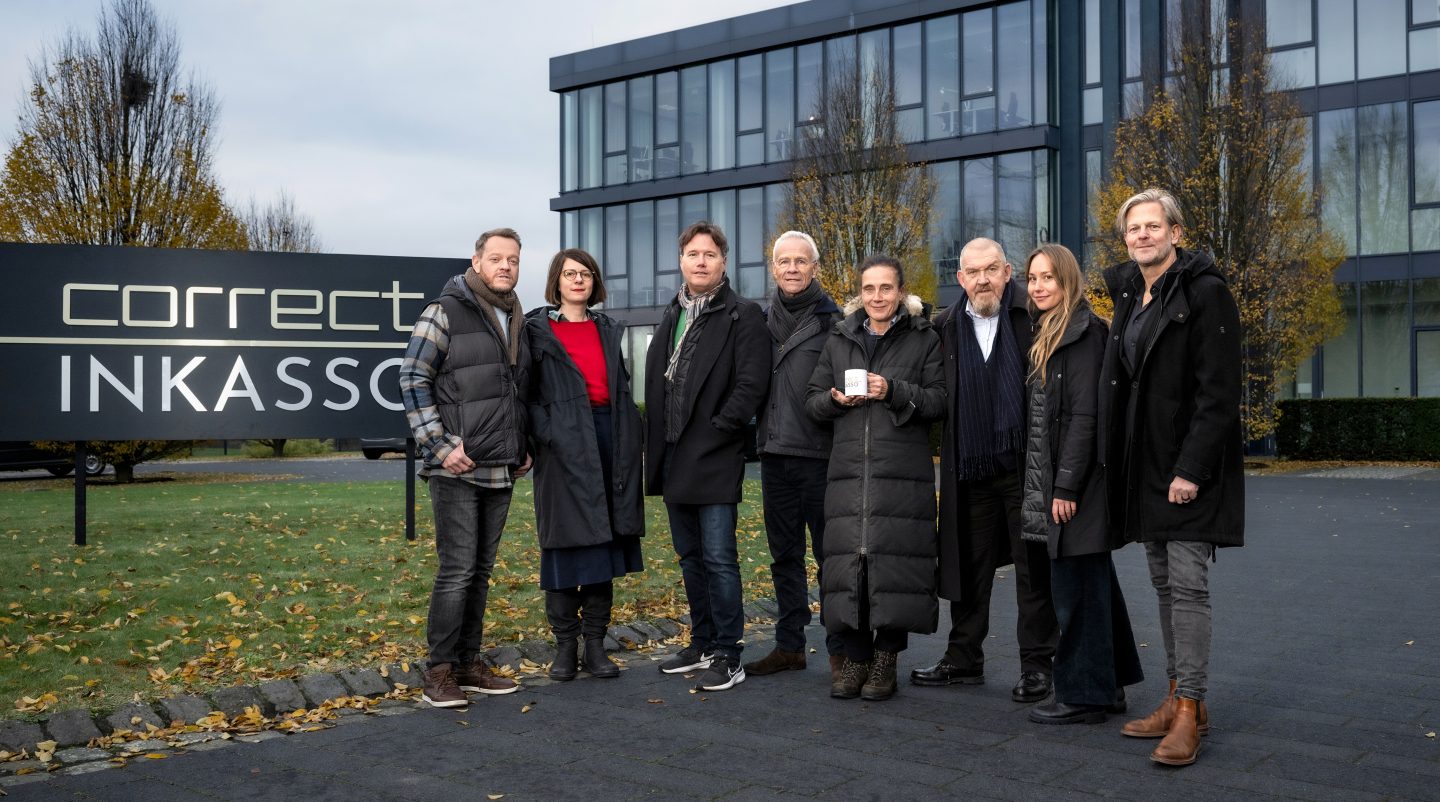 Gruppenfoto beim Drehstart des „Tatort: Restschuld“ vor dem Gebäude der fiktiven Firma Correct Inkasso. Abgebildet sind (v.l.n.r.): Roland Riebeling (Norbert Jütte), Karlotta Ehrenberg (Autorin), Götz Bolten (Redaktion/WDR), Klaus J. Behrendt (Max Ballauf), Claudia Garde (Regie), Dietmar Bär (Freddy Schenk), Lena Katharina Krause (Kamera) und Jan Kruse (Produzent/Bavaria Fiction). Die Szene zeigt die Hauptverantwortlichen des Projekts in entspannter Atmosphäre vor dem Firmenschild.