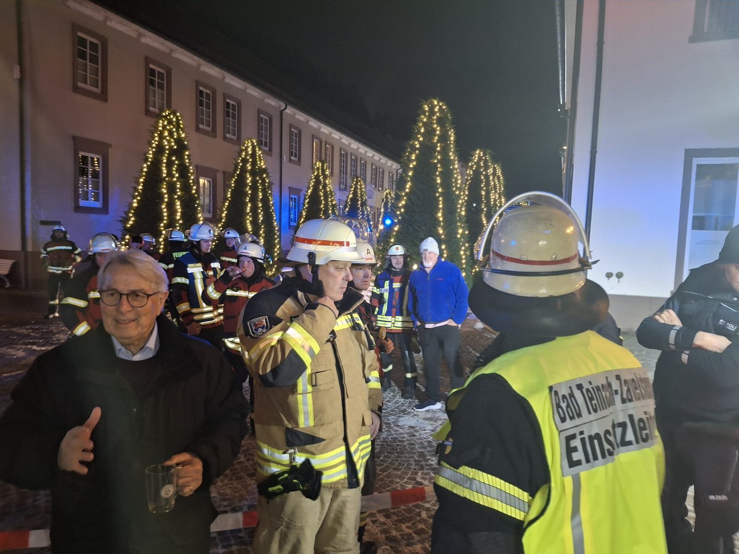 Einsatzleitung der Feuerwehr Bad Teinach-Zavelstein im Gespräch mit dem Hotelbesitzer vor dem evakuierten Gebäude, im Hintergrund Feuerwehrkräfte und weihnachtlich beleuchtete Bäume.