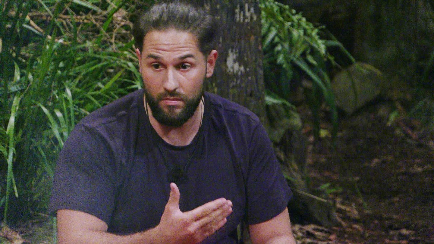 Maurice Dziwak sitzt mit ernstem Blick im Dschungelcamp und gestikuliert mit der Hand, während er sich gegen die Kritik von Jürgen verteidigt. Umgeben von dichtem Dschungelgrün wirkt er sichtlich getroffen von der Situation.