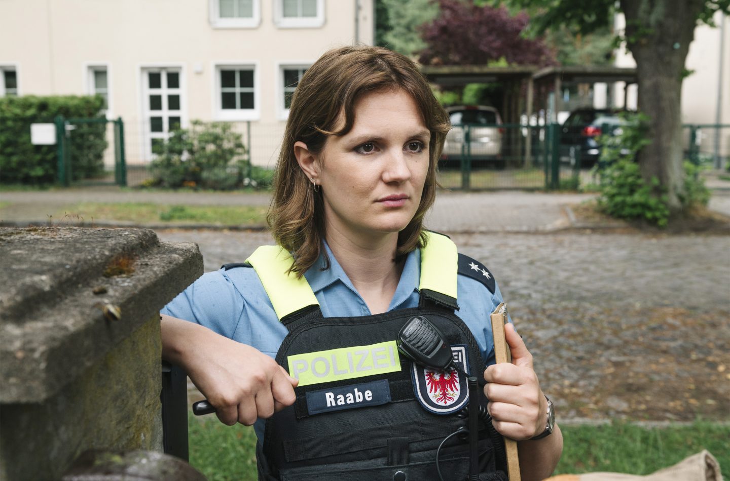 Tonja Raabe (Alina Stiegler) in Polizeiuniform mit Schutzweste steht vor einem Wohnhaus und hält Vermisstenanzeigen in der Hand. Ihr Gesichtsausdruck wirkt entschlossen und nachdenklich. Die Umgebung zeigt eine gepflasterte Straße mit geparkten Autos und einen Vorgarten, was auf eine Wohngegend hinweist.