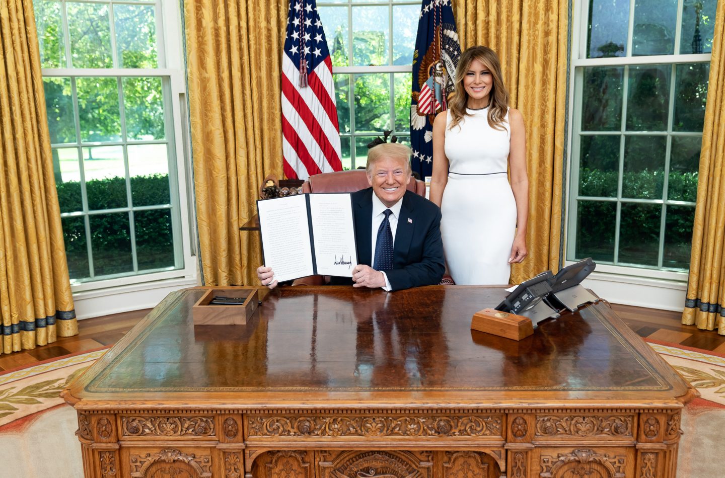 Donald Trump sitzt am Schreibtisch im Oval Office und hält ein unterzeichnetes Dokument in die Kamera. Neben ihm steht Melania Trump in einem weißen Kleid. Im Hintergrund sind die US-Flagge und große Fenster mit Blick ins Grüne zu sehen.