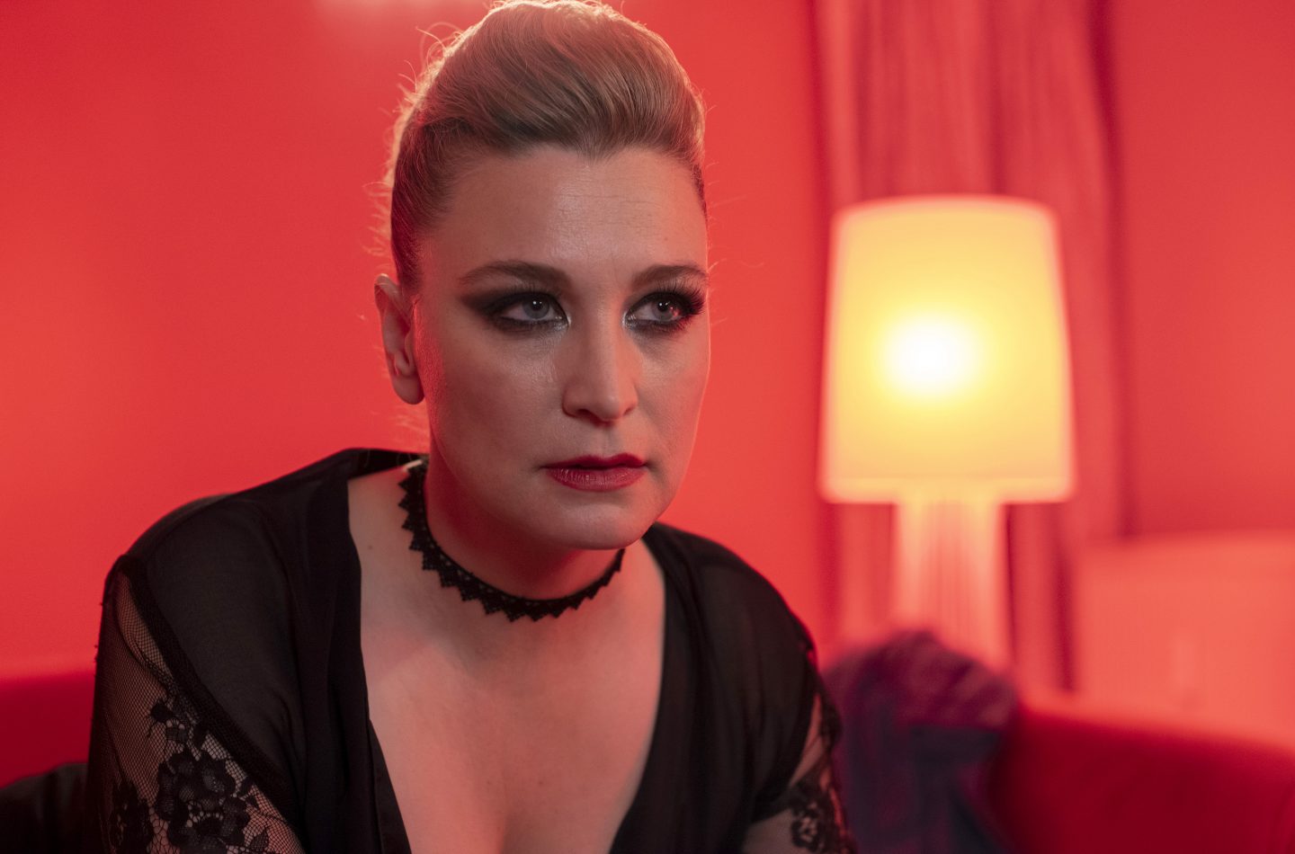 Anna König in der Rolle von Andrea Gonzor sitzt in einem rot ausgeleuchteten Raum, der durch eine warme Lichtquelle im Hintergrund belebt wird. Ihr ernster Ausdruck und das elegante schwarze Outfit mit Spitzenakzenten unterstreichen eine geheimnisvolle und intensive Atmosphäre.