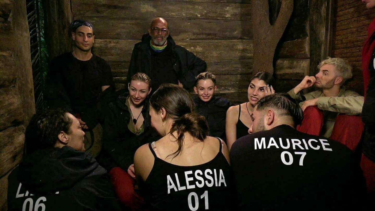 Die Dschungelcamp-Teilnehmer, darunter Lilly Becker, Timur Ülker, Anna-Carina Woitschack, Pierre Sanoussi-Bliss, Alessia Herren, Edith Stehfest, Yeliz Koc, Maurice Dziwak und Sam Dylan, sitzen eng gedrängt im Dschungeltelefon. Sie haben Schutz vor dem Starkregen und den Blitzen gesucht. Alessia und Maurice sind mit ihren Namen und Nummern auf den Rücken gekennzeichnet, während die Gruppe sichtlich durchnässt, aber in aufgelockerter Stimmung ist. Die rustikale Holzwand und der gedämpfte Lichtschein schaffen eine Atmosphäre des Zusammenhalts inmitten der Naturgewalten.