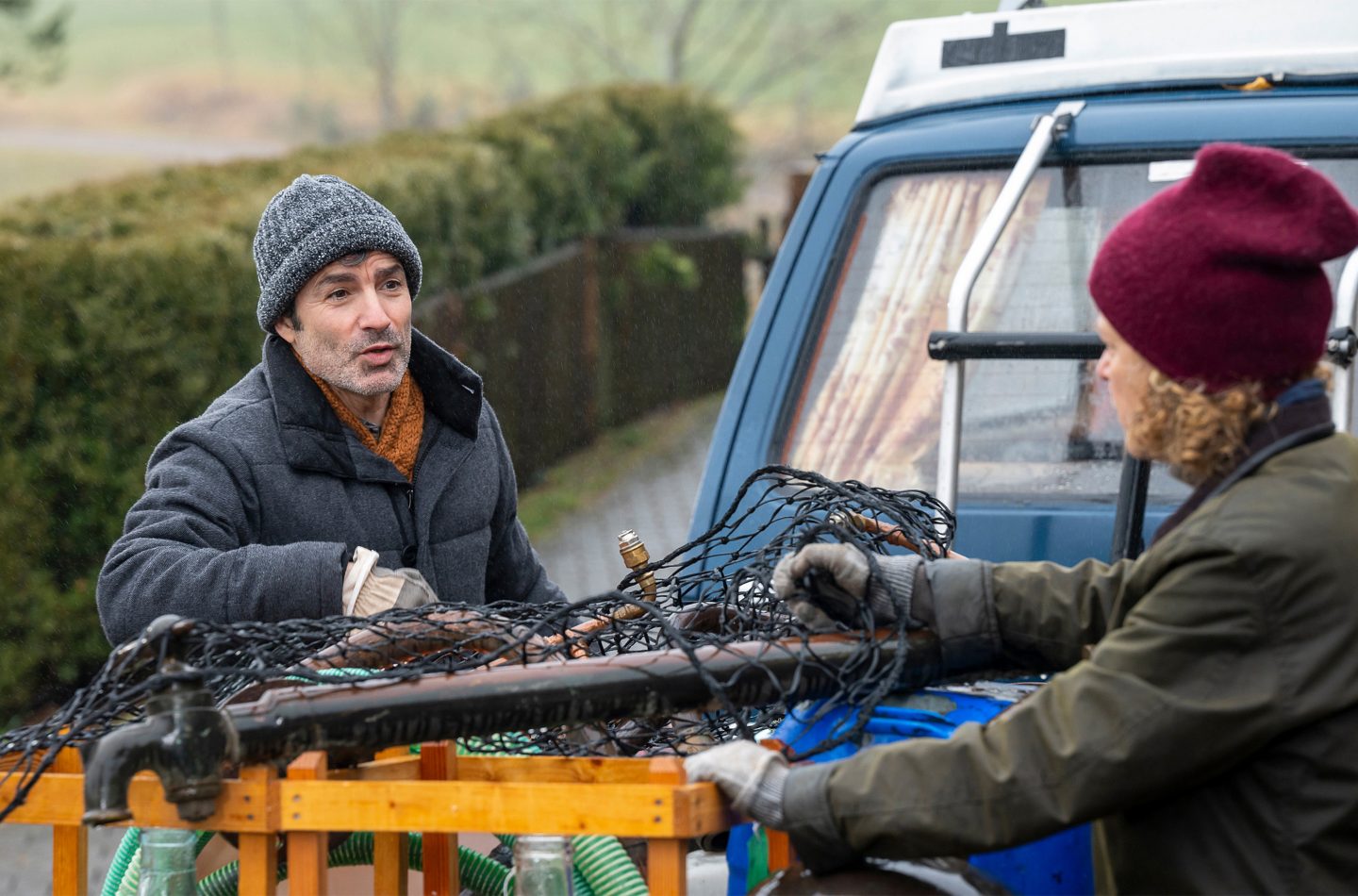 Marco Canadea als Manuel und Christiane Brammer als Bea stehen draußen vor einem blauen Transporter und diskutieren, während Manuel eine Destille in den Händen hält. Beide tragen warme Winterkleidung, und die Szene spielt an einem regnerischen Tag