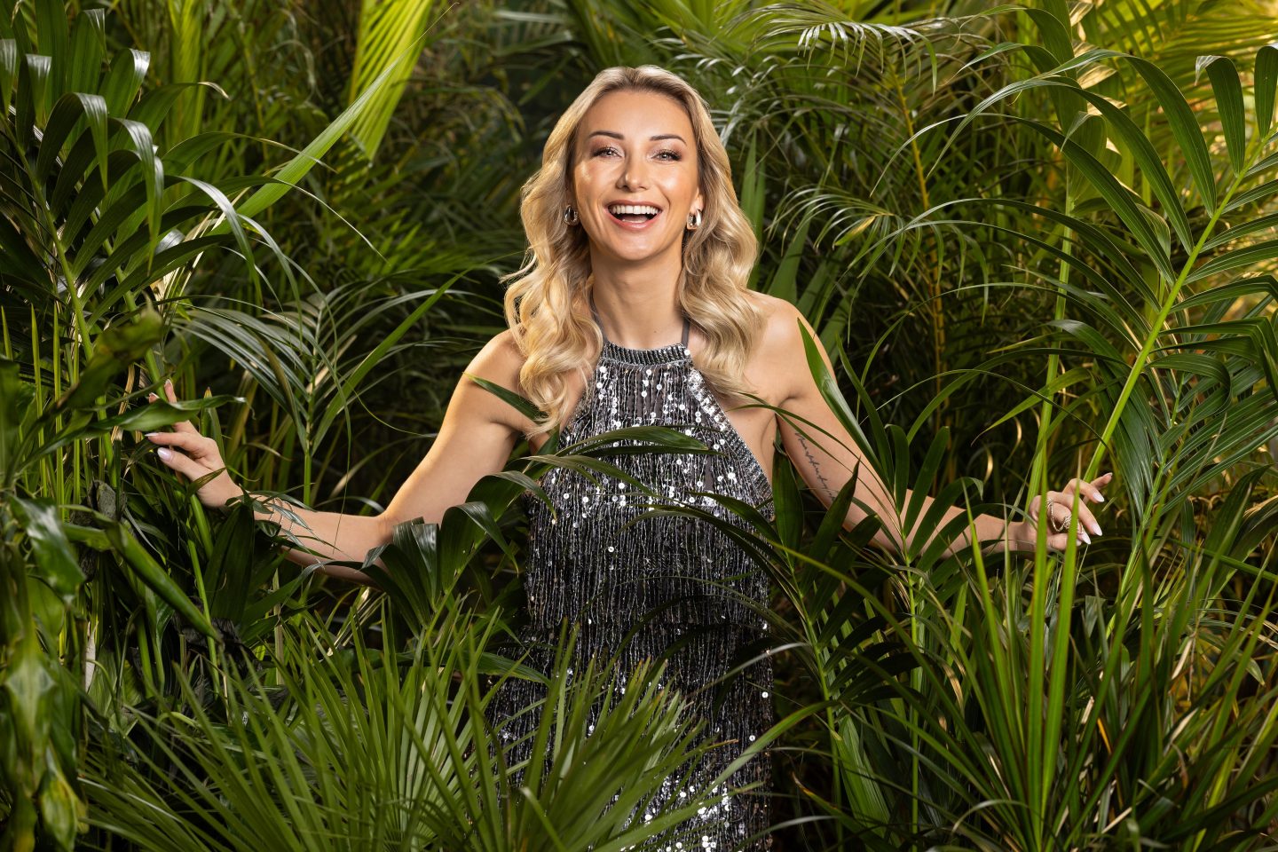 Anna-Carina Woitschack, strahlend in einem funkelnden silbernen Kleid, mit blonden Locken und einem breiten Lächeln, umgeben von tropischen Pflanzen im Dschungel.







