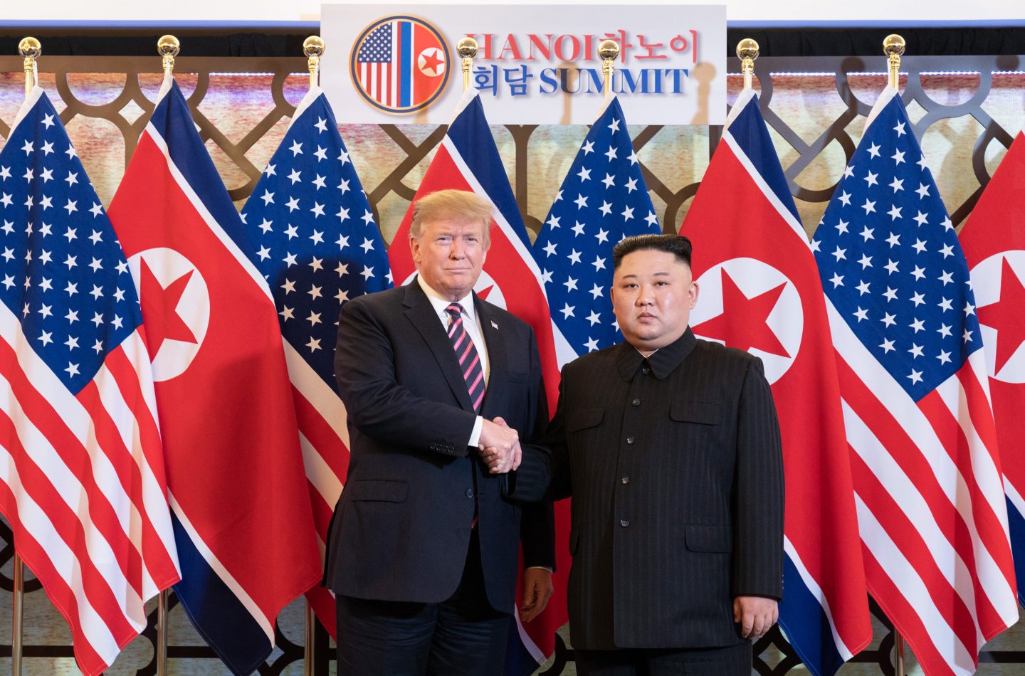Donald Trump und Kim Jong-un stehen vor einer Kulisse mit USA- und Nordkorea-Flaggen und schütteln sich die Hände während des Hanoi-Gipfels. Im Hintergrund ist ein Schild mit der Aufschrift 'HANOI SUMMIT' sichtbar.