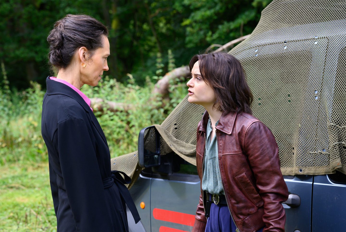 Mutter Béatrice Radek (Sabine Timoteo, links) und ihre Tochter Carla Radek (Lena Urzendowsky, rechts) stehen sich in einer angespannten Konfrontation gegenüber. Béatrice, elegant gekleidet in einem schwarzen Mantel mit rosa Kragen, blickt ernst und distanziert auf ihre Tochter. Carla, in einer braunen Lederjacke und blauer Hose, wirkt aufgebracht und konfrontativ. Im Hintergrund ist ein mit Tarnnetz bedecktes Fahrzeug sowie dichtes grünes Gebüsch zu sehen, was auf eine ländliche Umgebung hindeutet.